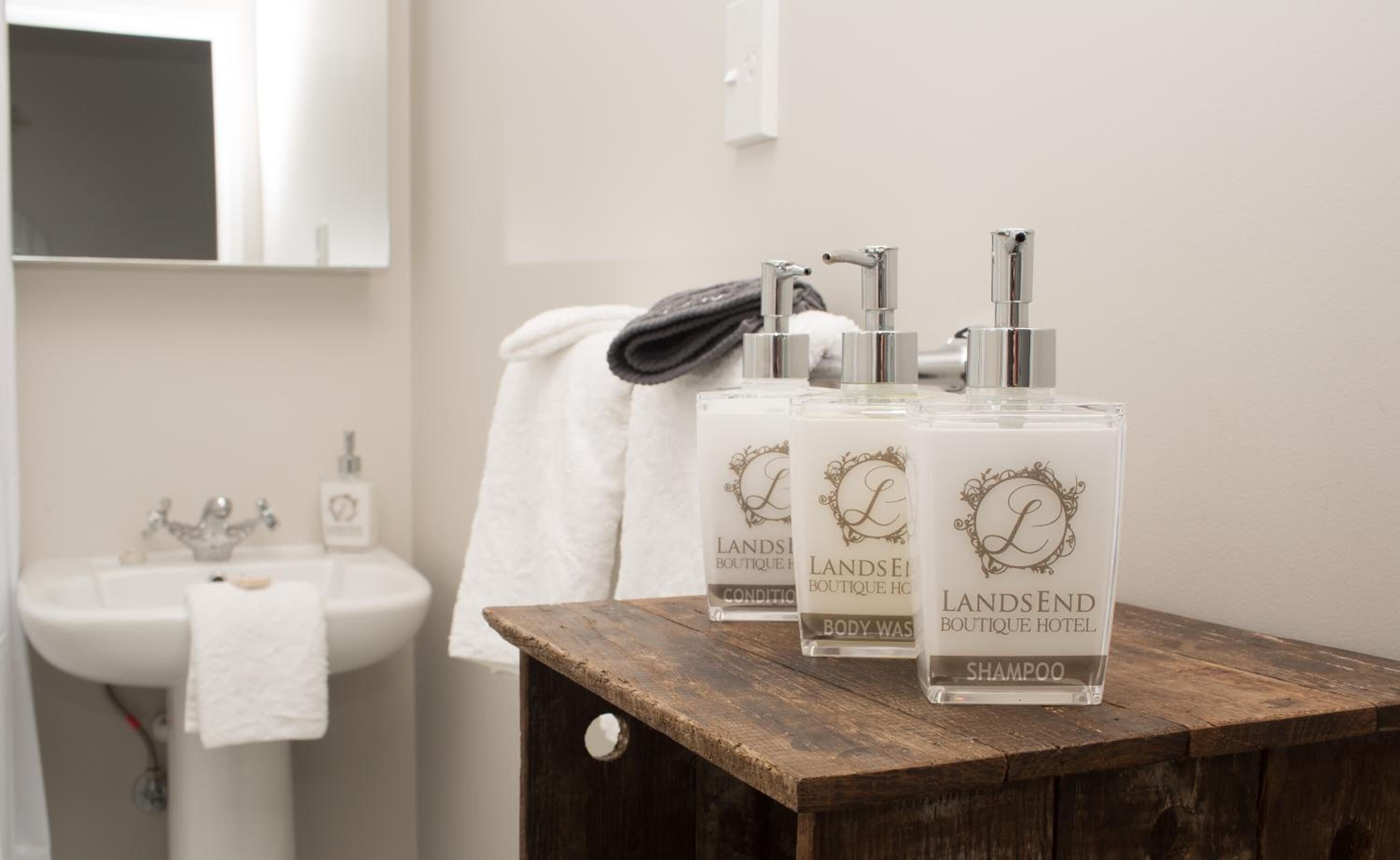 Tres botellas de champú sobre una superficie de madera en un baño con lavamanos y toalla.