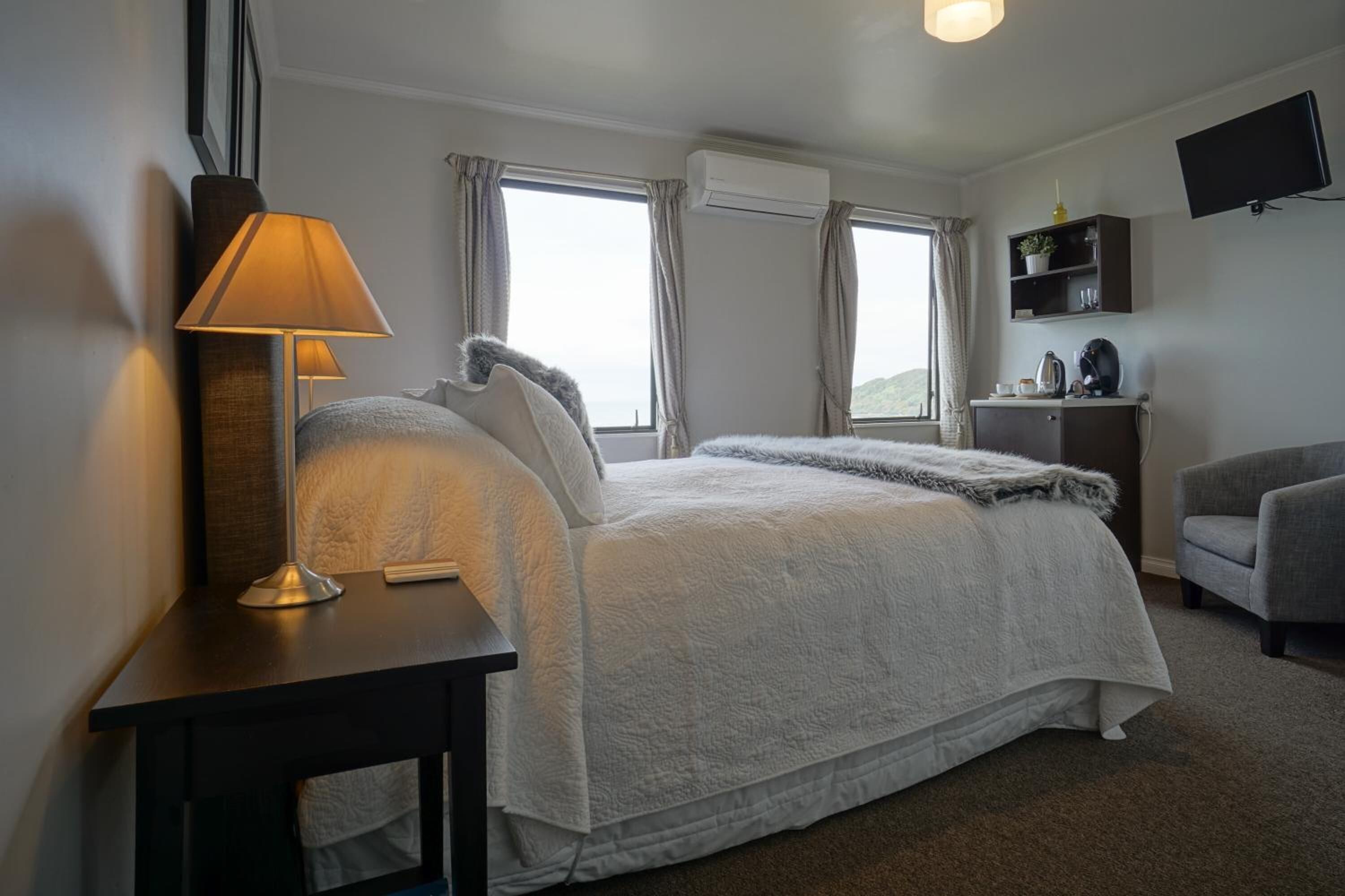 Un dormitorio con una cama grande, una mesita de noche con lámpara, una silla, un televisor en la pared y un estante con objetos.