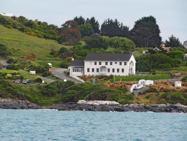 A white building with a steep roof, surrounded by lush greenery and a rocky shoreline.