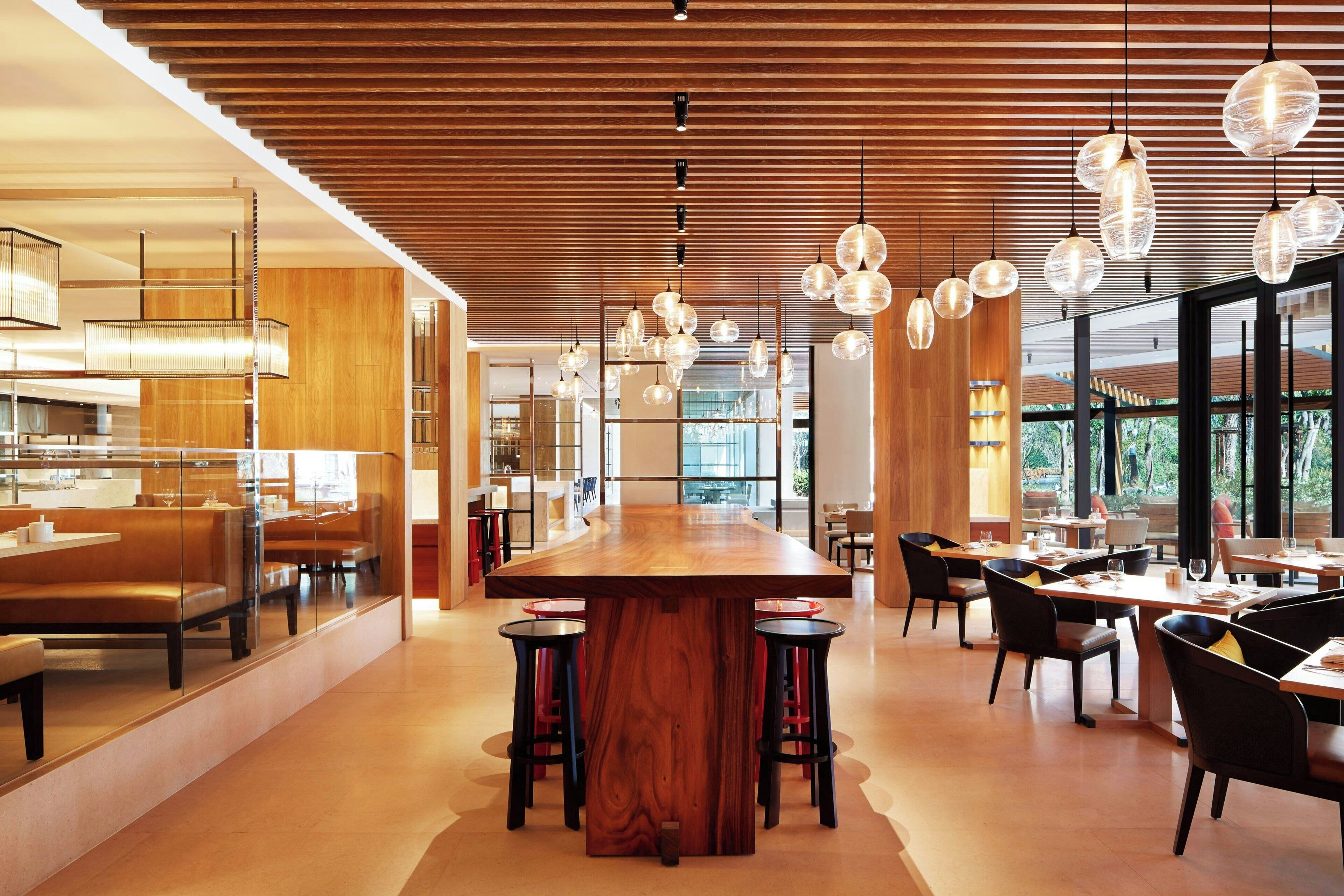 A modern restaurant with a central wooden bar, multiple hanging pendant lights, and a spacious layout with high ceilings and large windows.