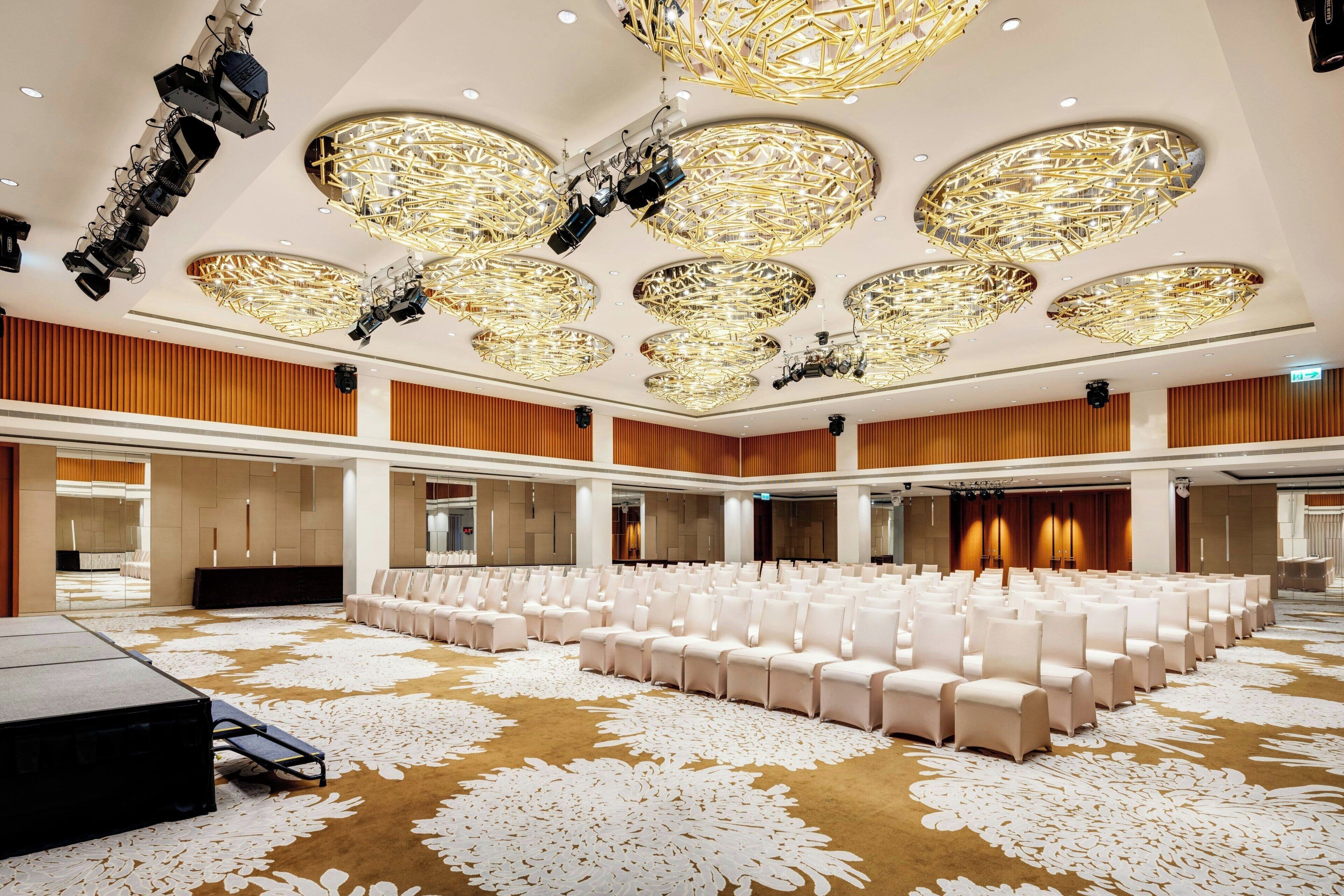 A spacious conference room with rows of white chairs, a large carpet with a floral pattern, and multiple ceiling lights with a unique design.
