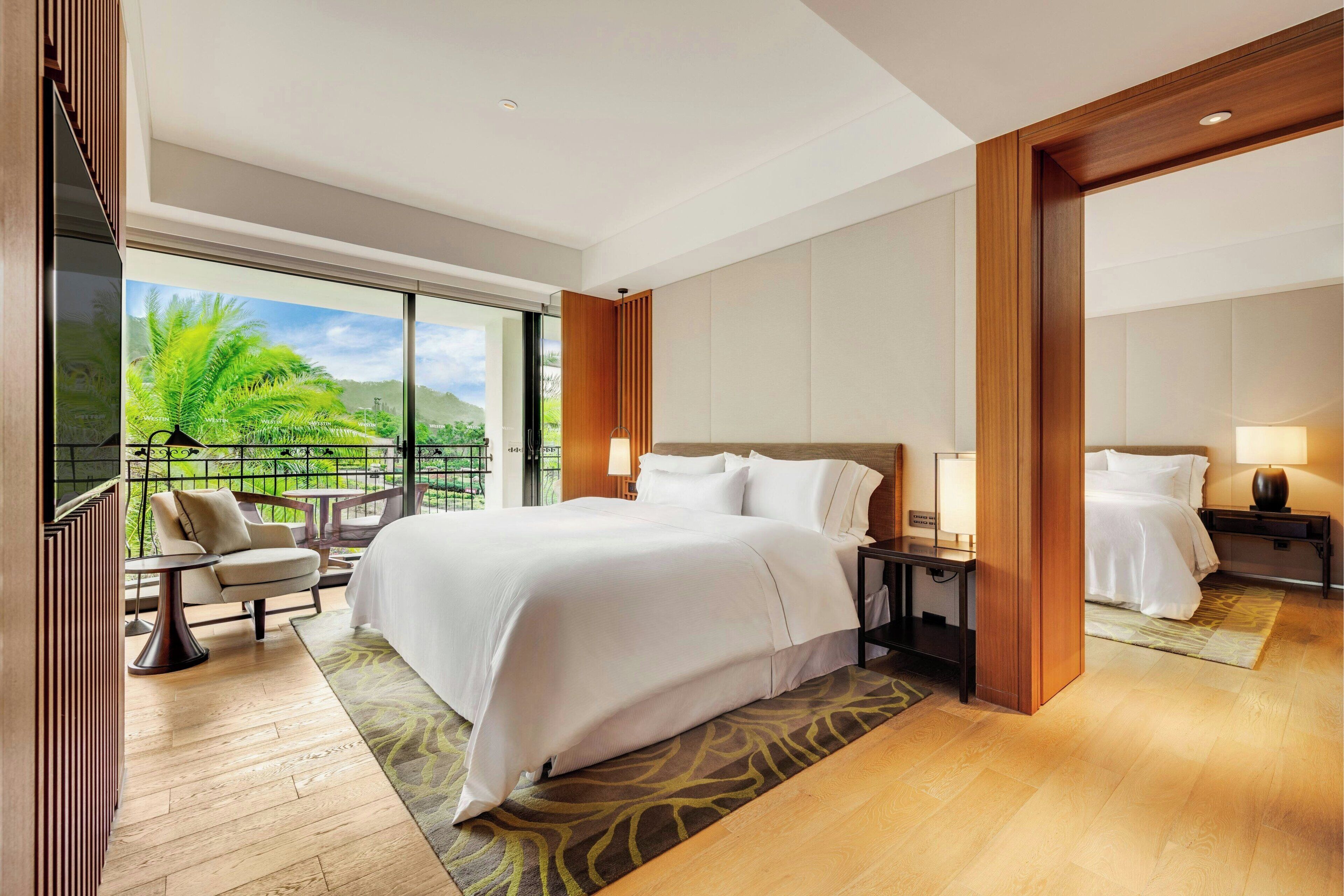 Habitación de hotel con una cama grande, una zona de estar con una silla y una mesita, una mesita de noche con lámpara, y vistas a zonas verdes a través de amplias ventanas.