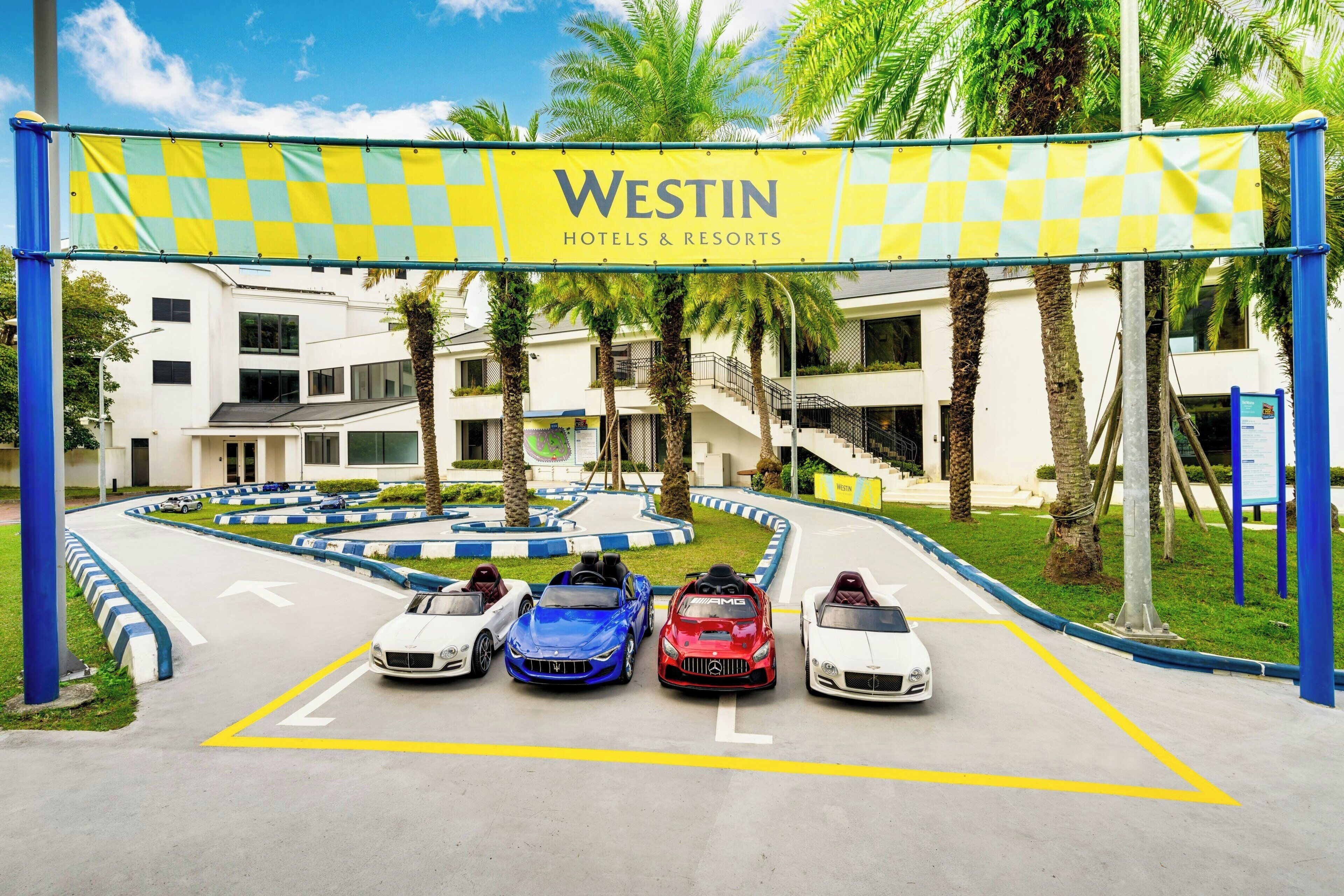 A Westin Hotels & Resorts entrance with a checkered banner, palm trees, and a parking area with four cars.