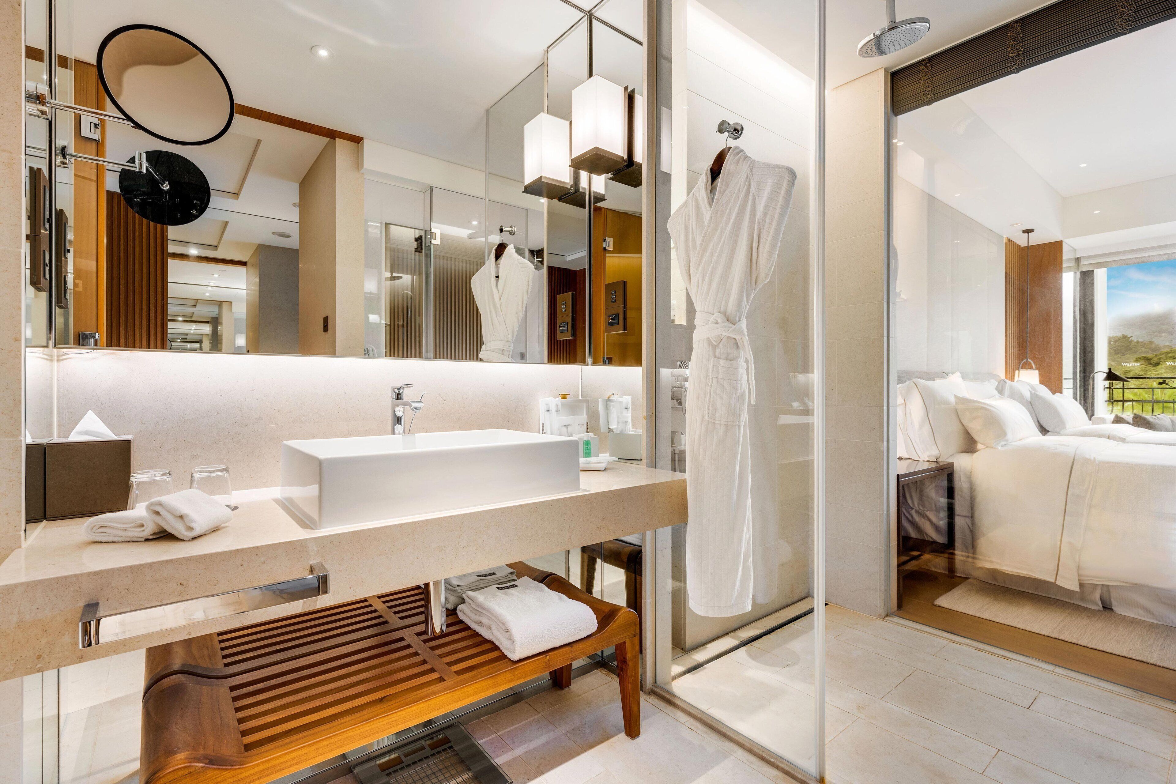 Une salle de bain moderne avec un grand miroir, un banc en bois, un lavabo blanc et un peignoir suspendu à un porte-serviettes.