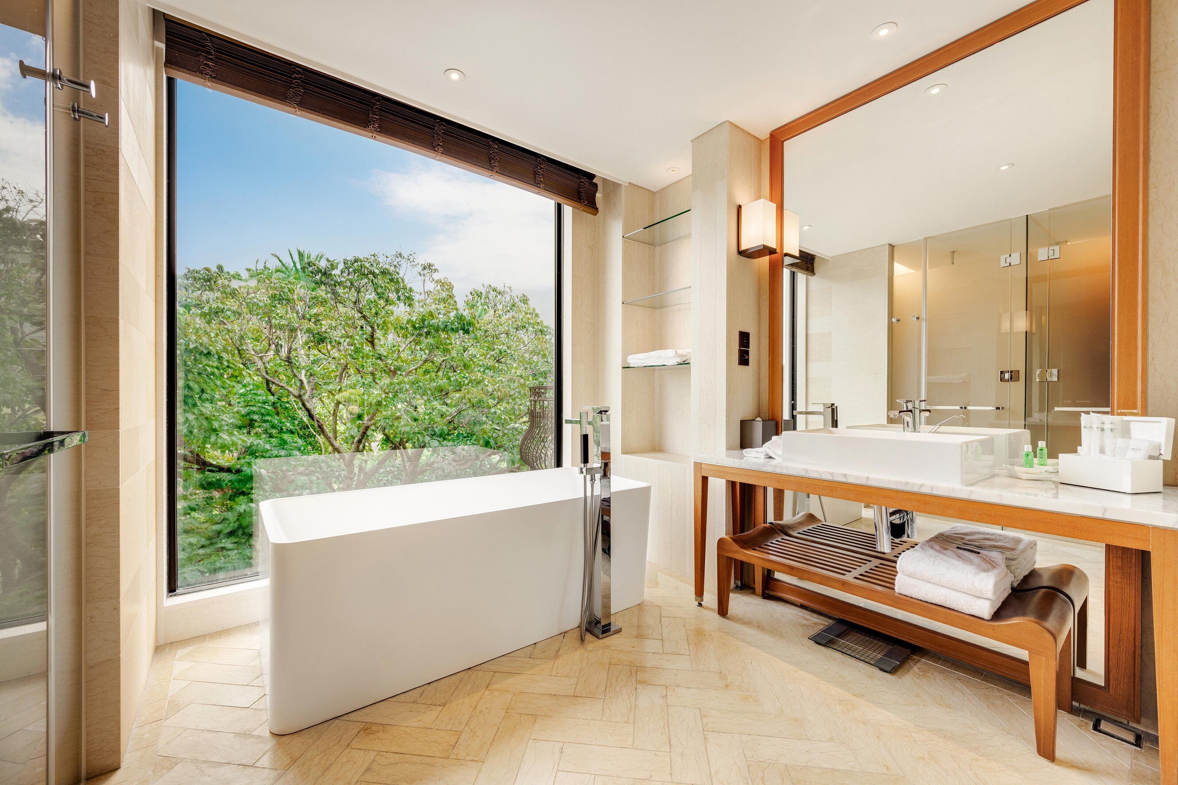 A modern bathroom with a large bathtub, a wooden bench, a large mirror, and a view of greenery outside.