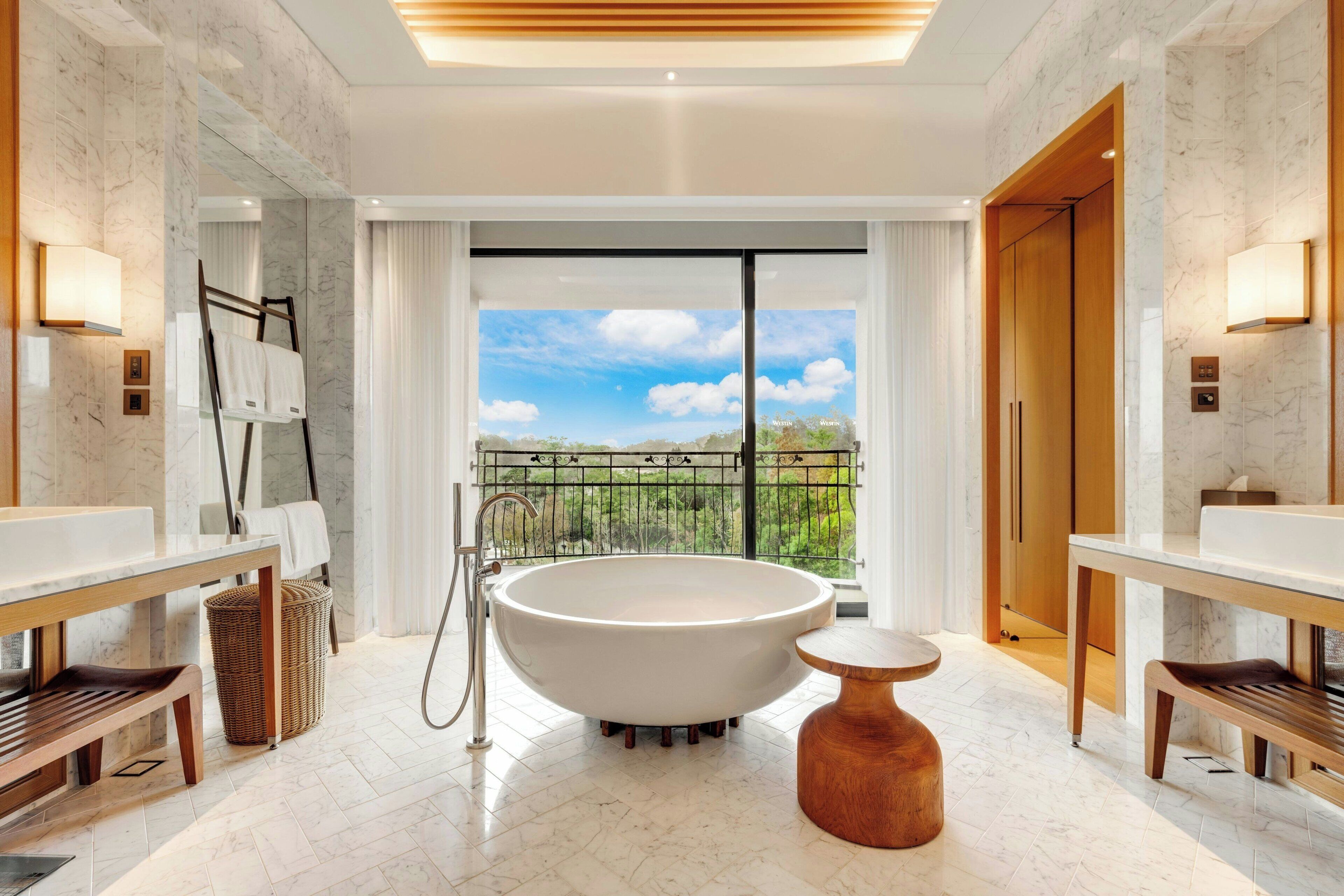 A modern bathroom with a freestanding bathtub, wooden benches, and a view of a green landscape through large windows.