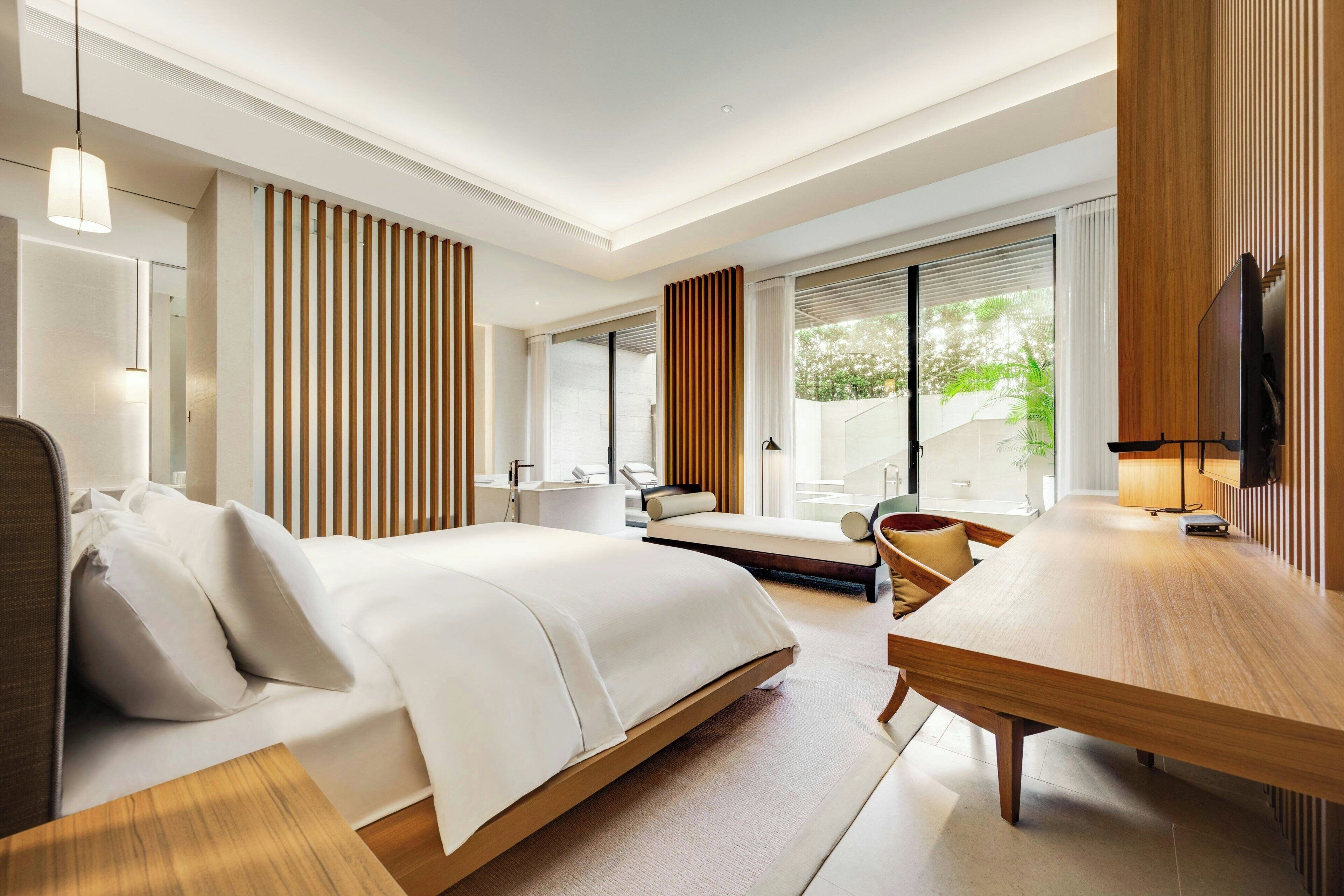 A modern hotel room with a large bed, a wooden desk, and a view of greenery outside.