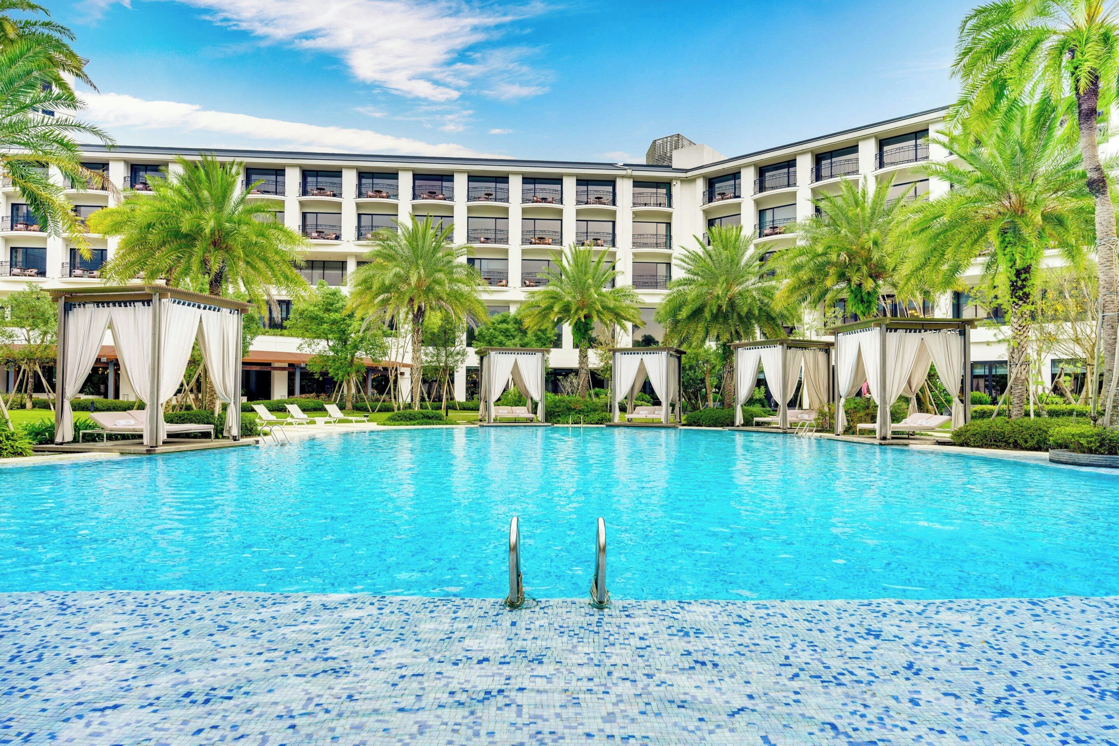 A large swimming pool with clear blue water, surrounded by palm trees and a multi-story building with balconies.