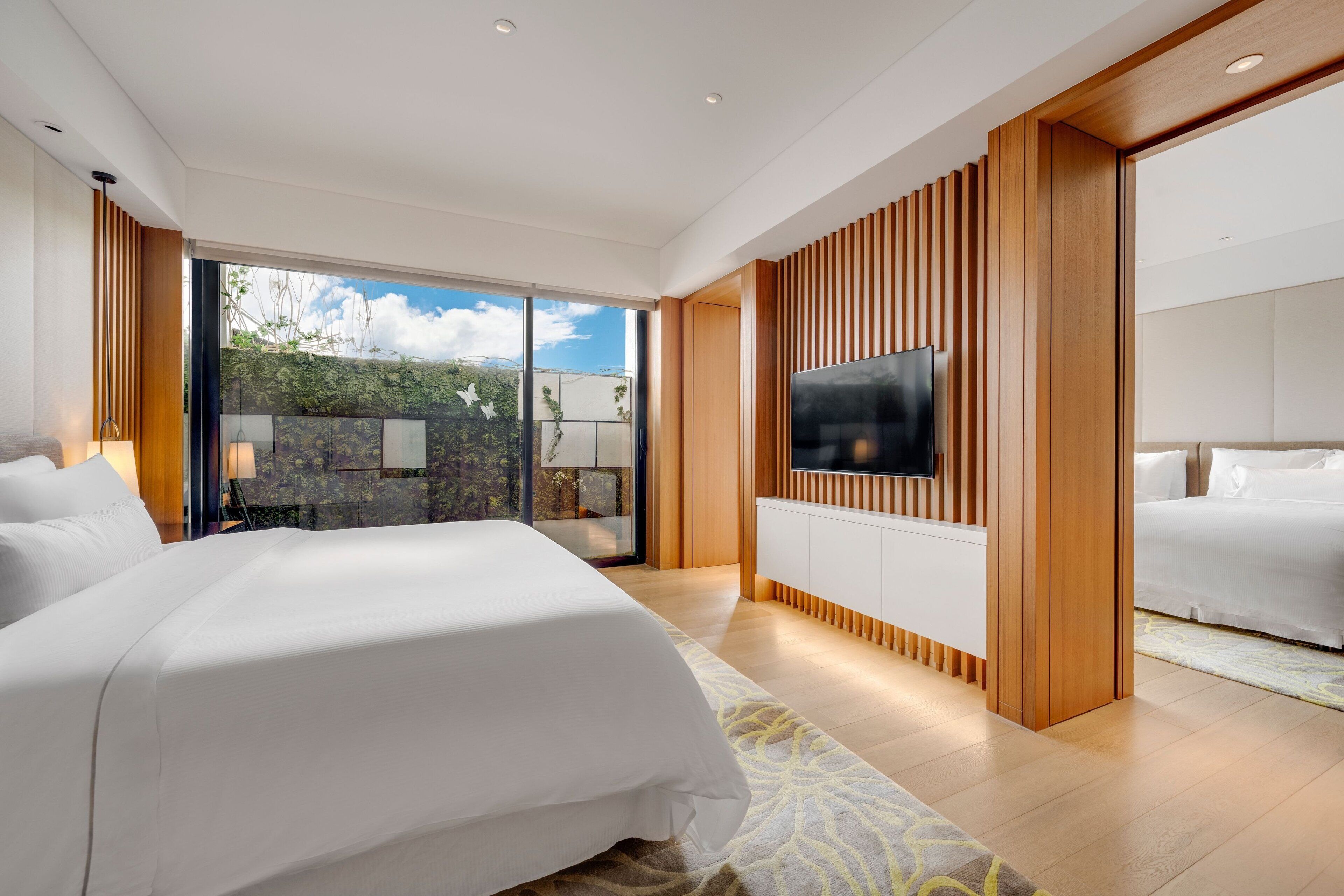 A modern hotel room with a large bed, a flat-screen TV mounted on a wooden panel, and a balcony with a view of greenery.