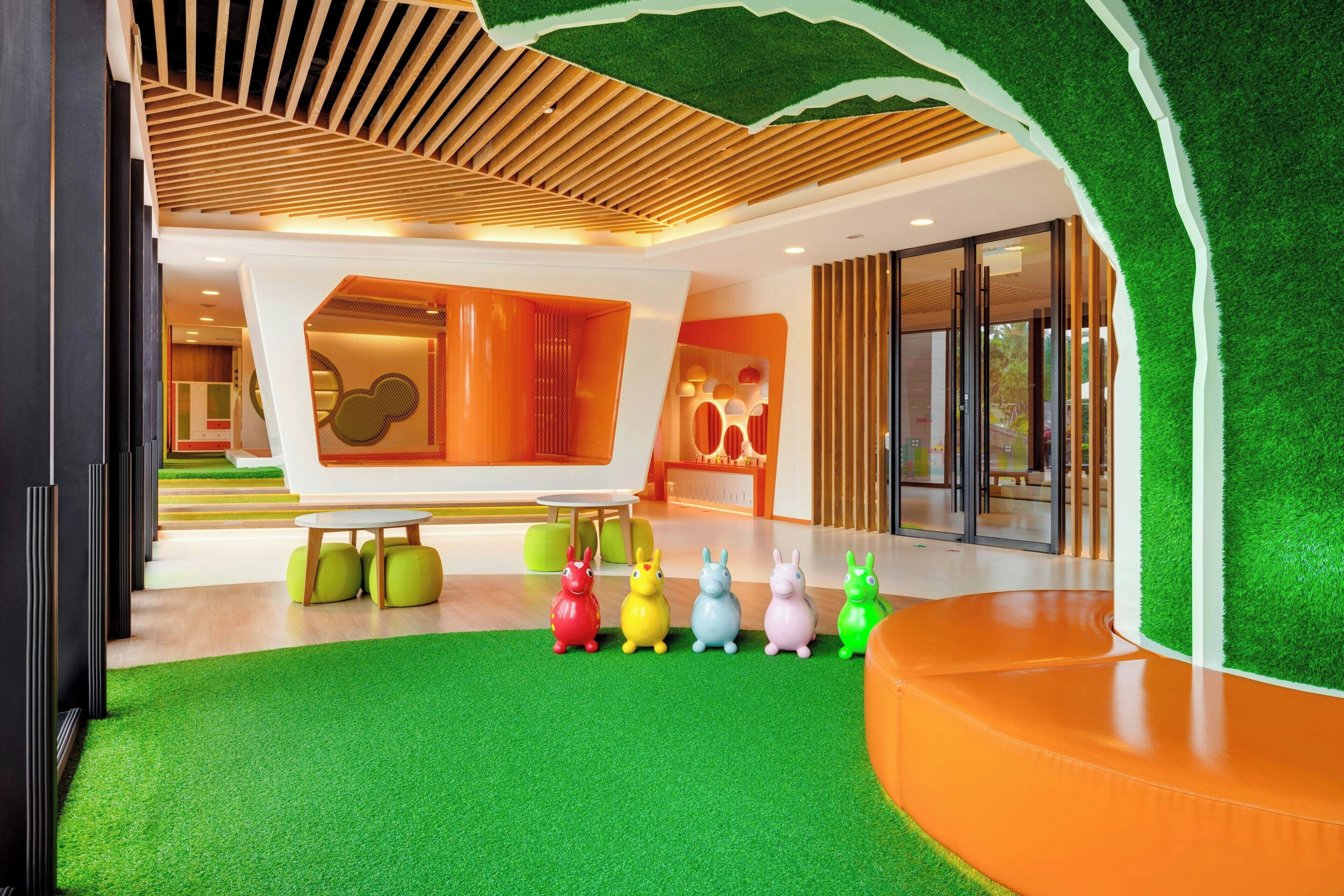 Um interior moderno com piso de grama sintética verde, tabouretos em formato de animais coloridos e teto de ripas de madeira.