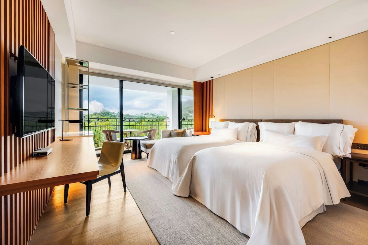 Ett modernt hotellrum med två sängar, ett skrivbord med en tv och en balkong med utsikt över den grönskande omgivningen.