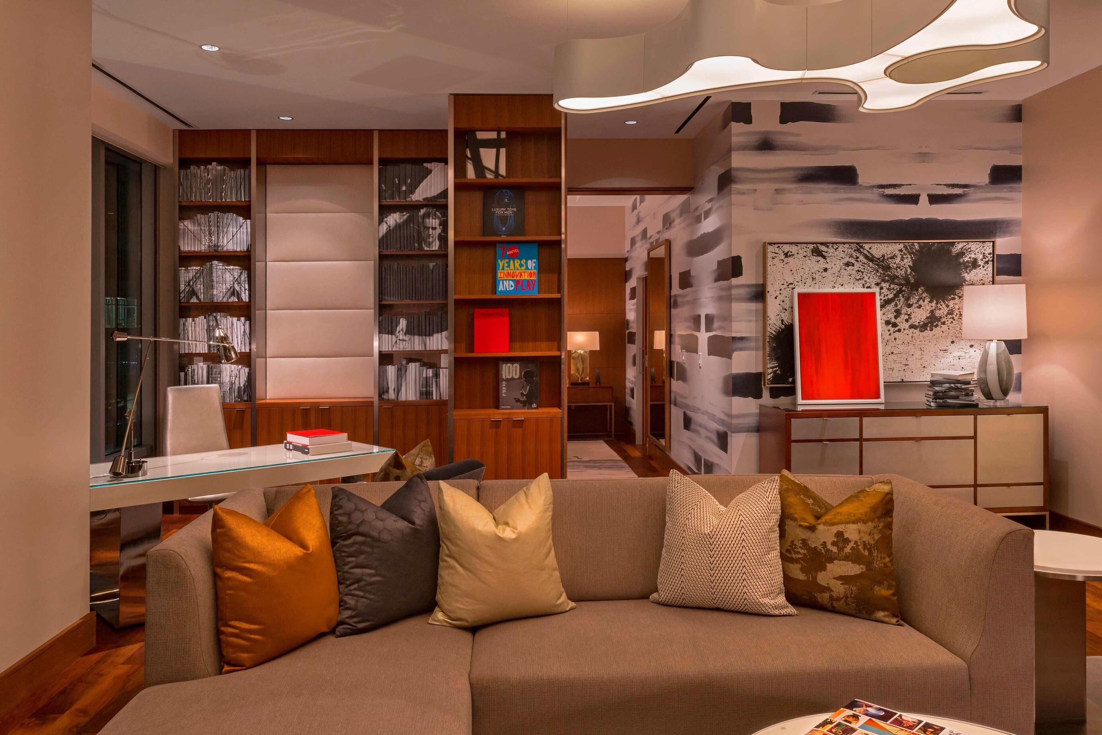 A modern living room with a glass coffee table, a beige sofa with patterned cushions, a bookshelf with various items, and a red accent wall.