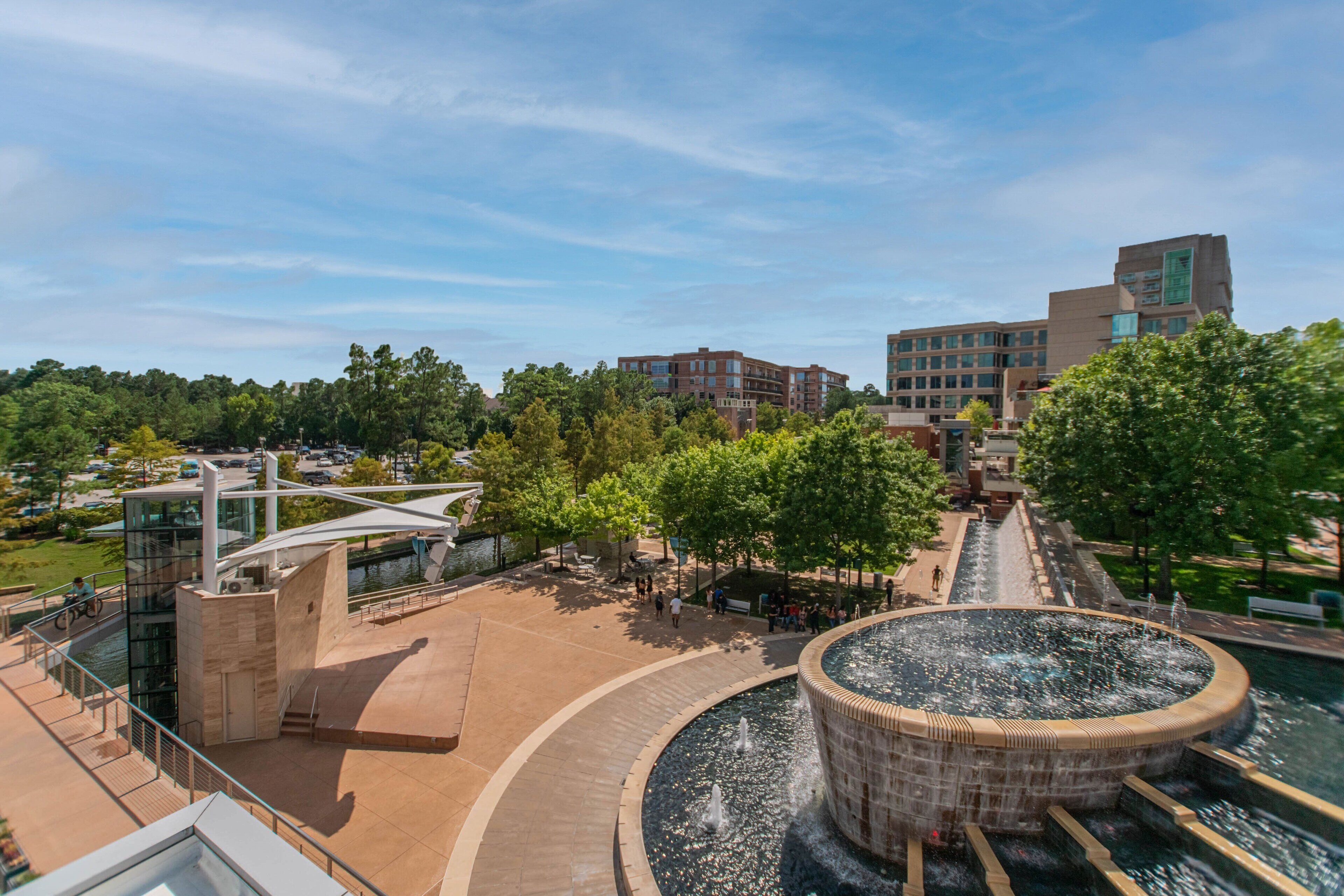 A modern building with a circular water feature in the center, surrounded by a plaza with benches and trees.