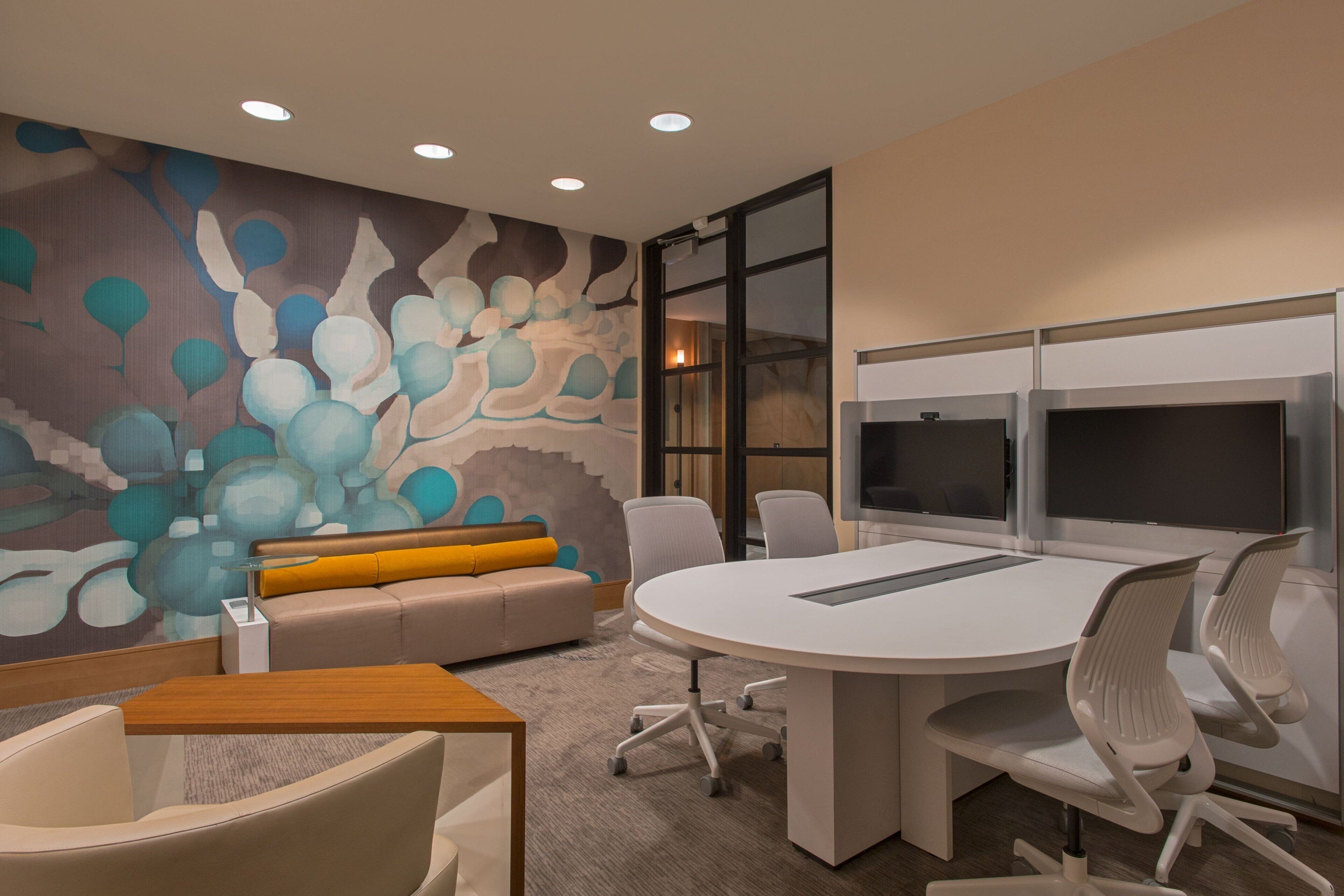A modern meeting room with a round table, white chairs, a sofa with a yellow cushion, and a colorful abstract mural on the wall.