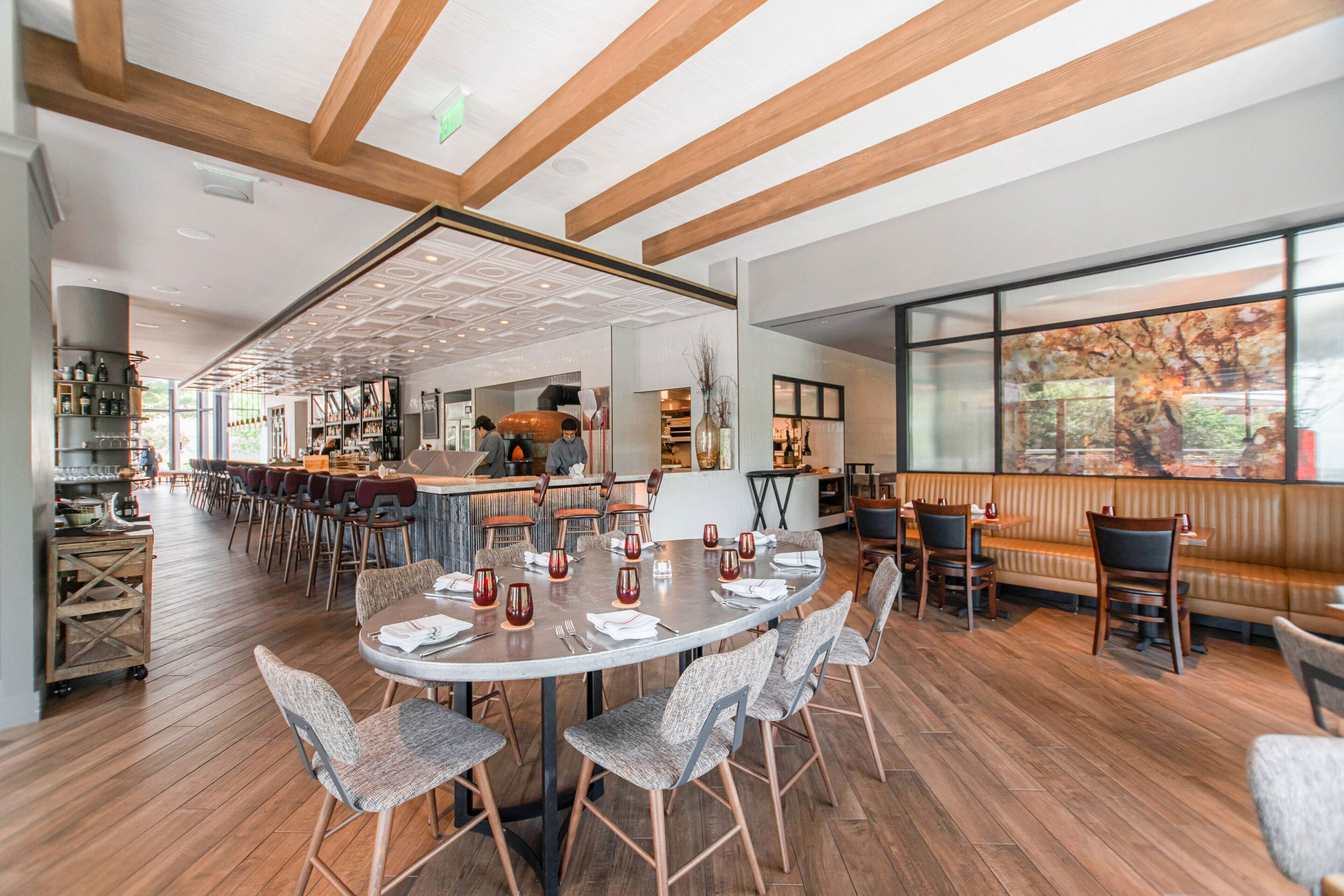 A spacious restaurant with wooden floors, a central dining area with round tables and chairs, and a bar area with stools.