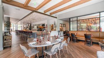 A spacious restaurant with wooden floors, a central dining area with round tables and chairs, and a bar area with stools.