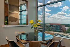 A round glass table with six chairs, a vase with yellow flowers, and a view of buildings through large windows.