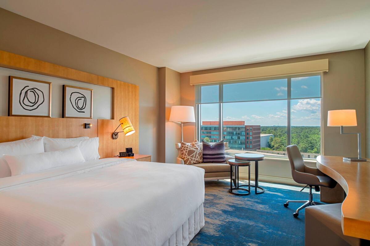 Habitación de hotel con una cama grande, dos lámparas de noche, un escritorio con una silla y vistas de la ciudad a través de amplios ventanales.