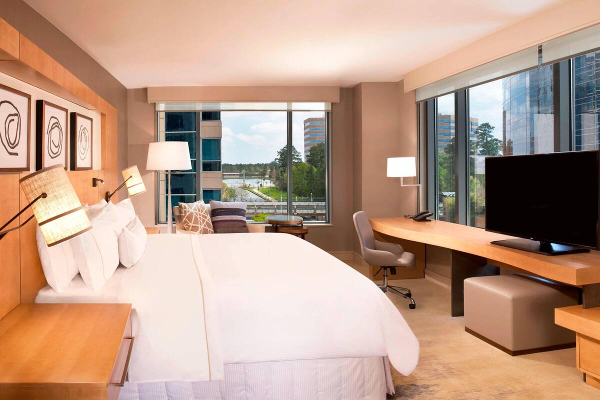 Habitación de hotel moderna con una cama grande, un escritorio con televisor de pantalla plana, una silla y vistas a un paisaje urbano a través de amplias ventanas.