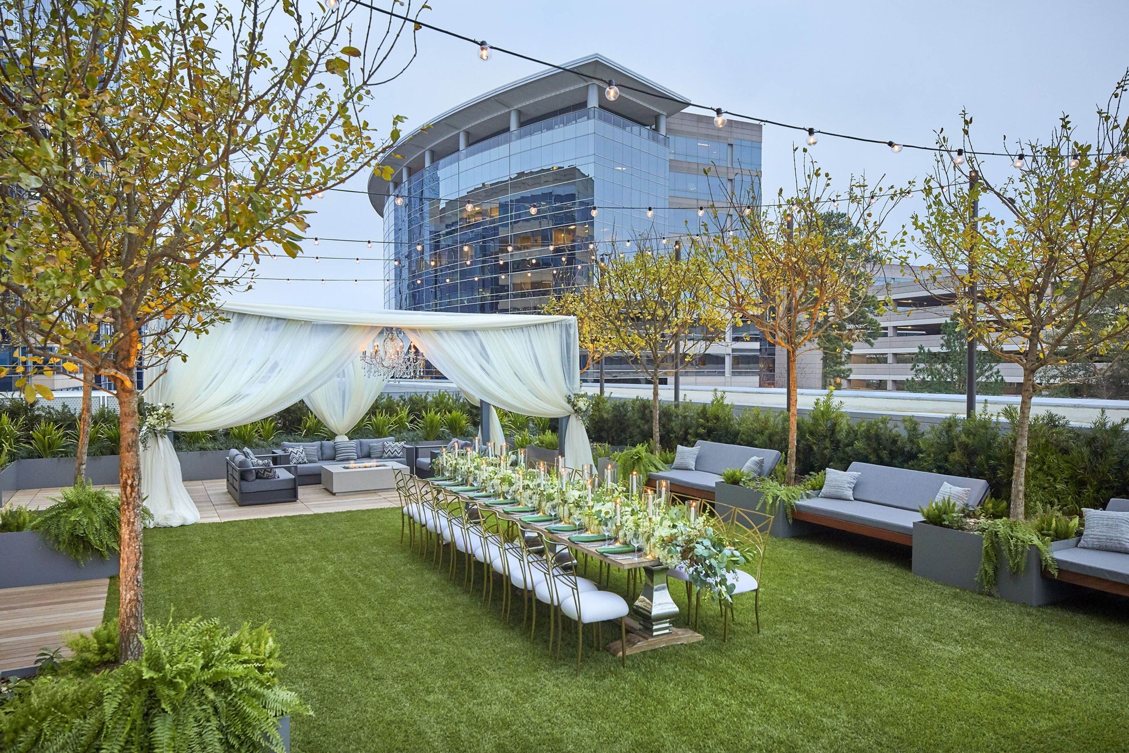 An outdoor dining area with a long table set for a meal, surrounded by seating and greenery.