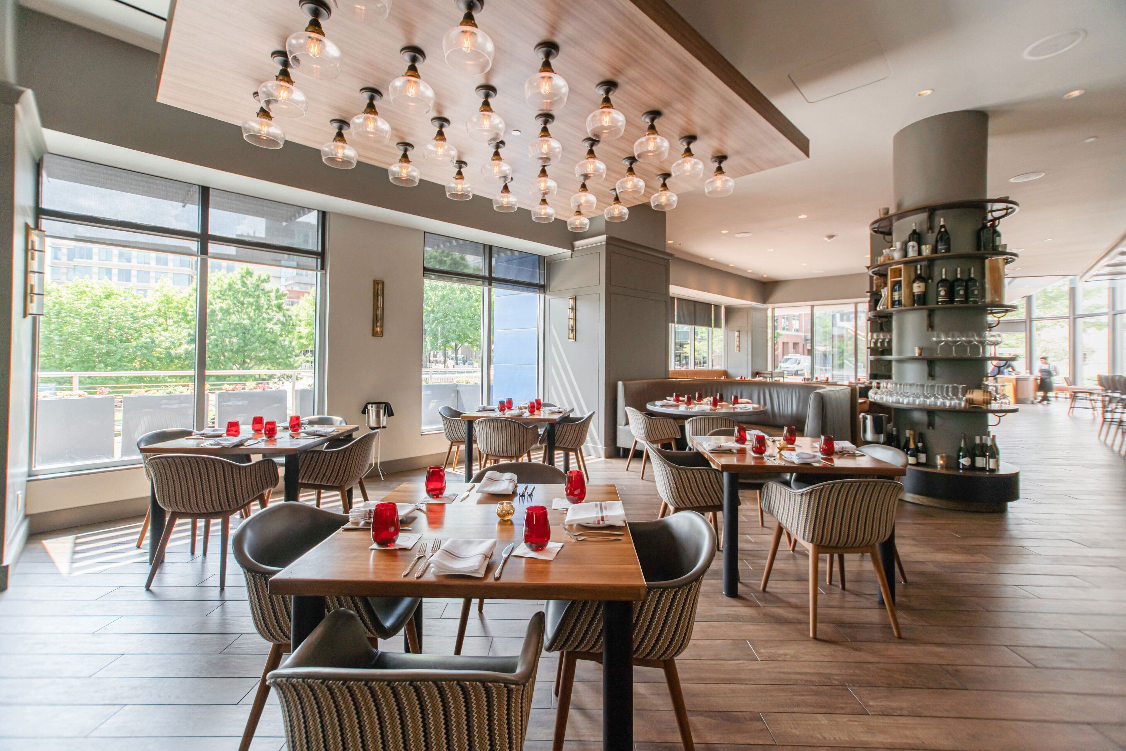 Un restaurante moderno con techos altos, vigas de madera a la vista y amplios ventanales que ofrecen vistas al exterior. Las mesas están dispuestas con copas de vino tinto y servilletas.