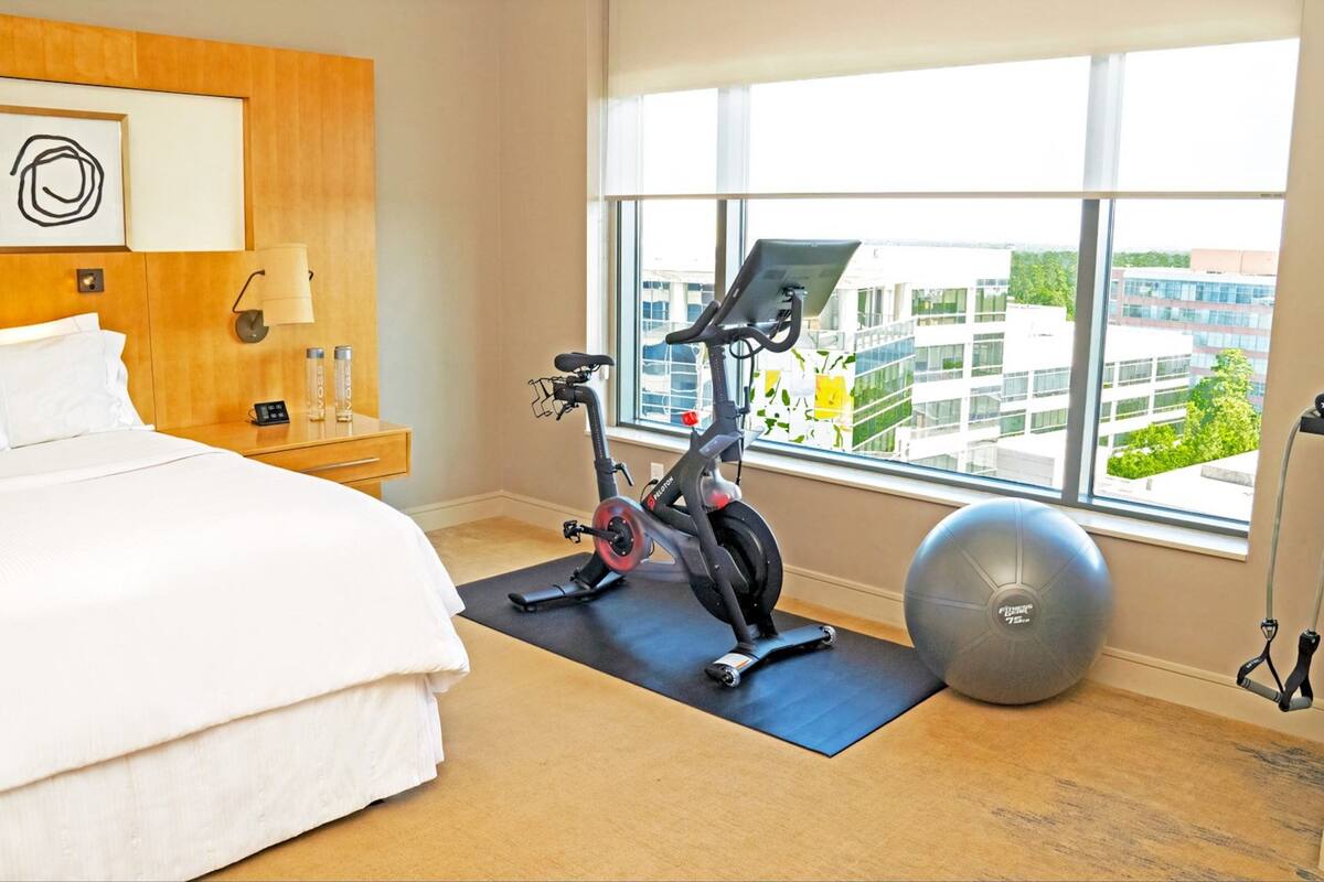 A hotel room with a bed, exercise bike, exercise ball, and a window with a city view.