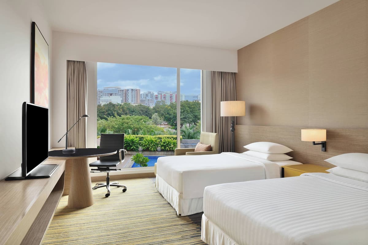 Ein Hotelzimmer mit zwei Betten, einem Schreibtisch mit Fernseher, einer Lampe und Blick auf Stadt und Grünflächen.