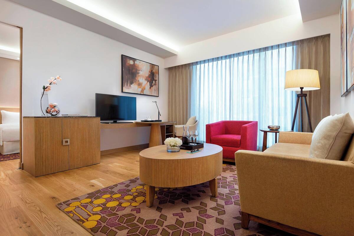Ein modernes Hotelzimmer mit Holzboden, einem runden Couchtisch, einem roten Sessel, einem beigen Sofa, einem Fernseher, einem Schreibtisch und einem großen Fenster mit Vorhängen.