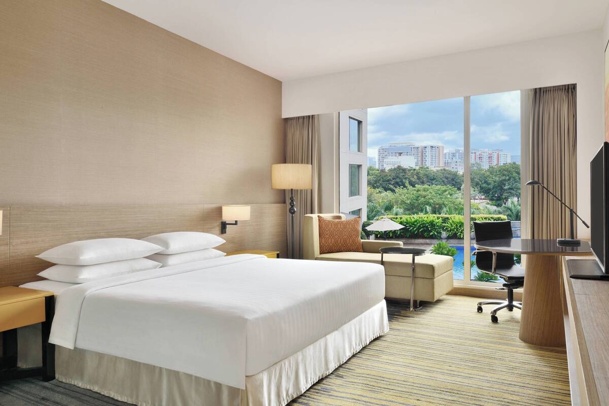 Ein Hotelzimmer mit einem großen Bett, einem Schreibtisch, einem Sessel, einem Fernseher und Blick auf Stadt und Grünflächen.