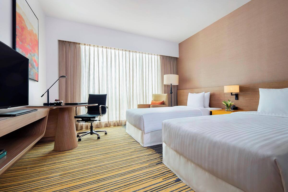 Ein Hotelzimmer mit zwei Betten, einem Schreibtisch, einem Fernseher und einem großen Fenster mit Vorhängen.