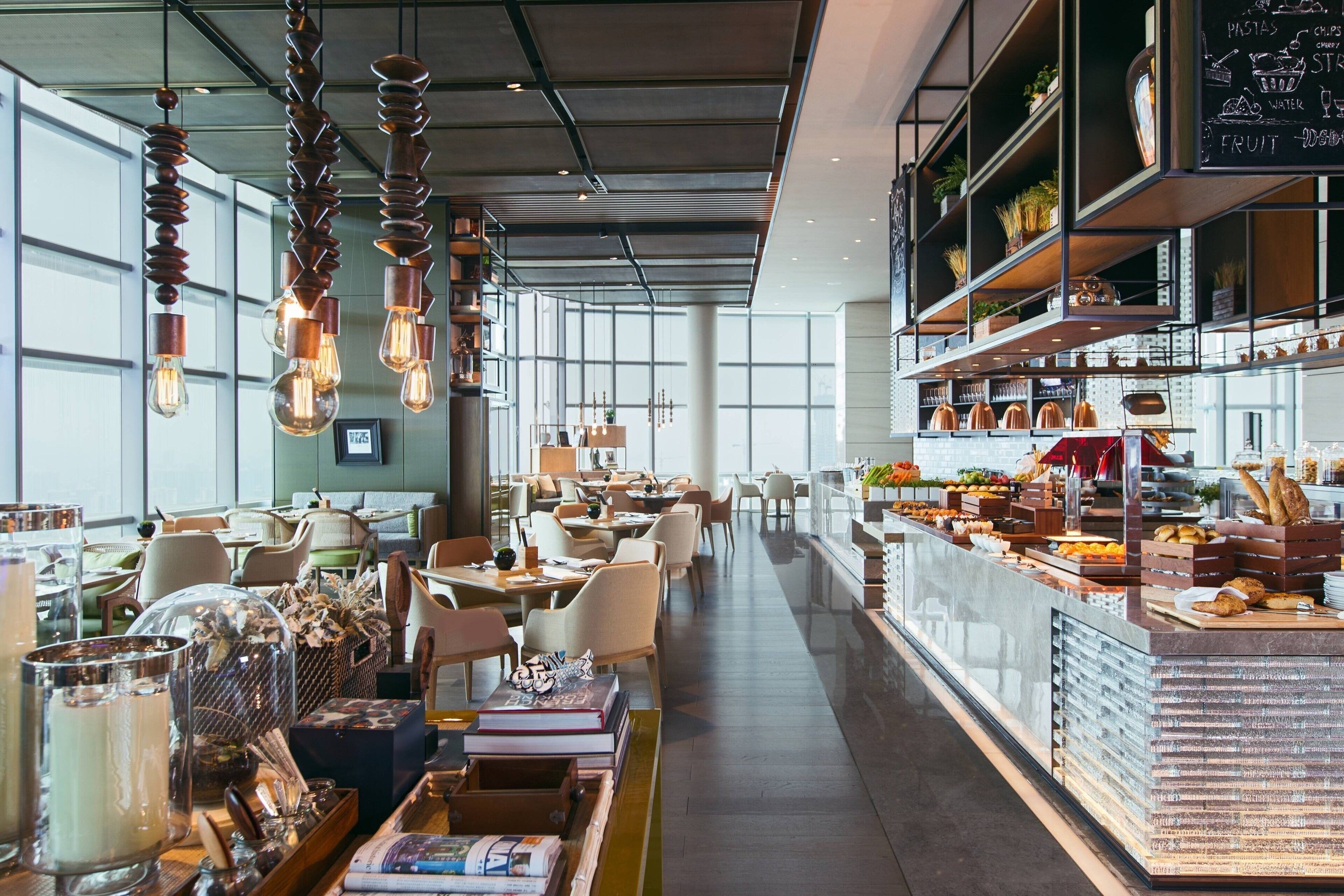 A modern cafe with a long counter displaying pastries and drinks, hanging light fixtures, and a view of the city through large windows.