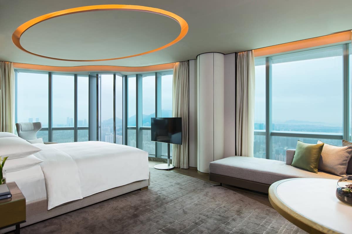 A modern hotel room with a large bed, a seating area, a flat-screen TV, and a large window offering a city view.