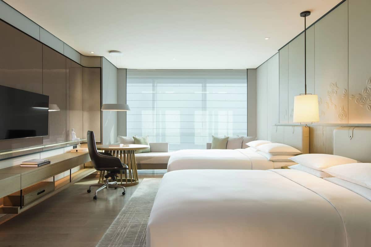 A modern hotel room with a large flat-screen TV, a desk with a chair, two beds, and a large window with a view.