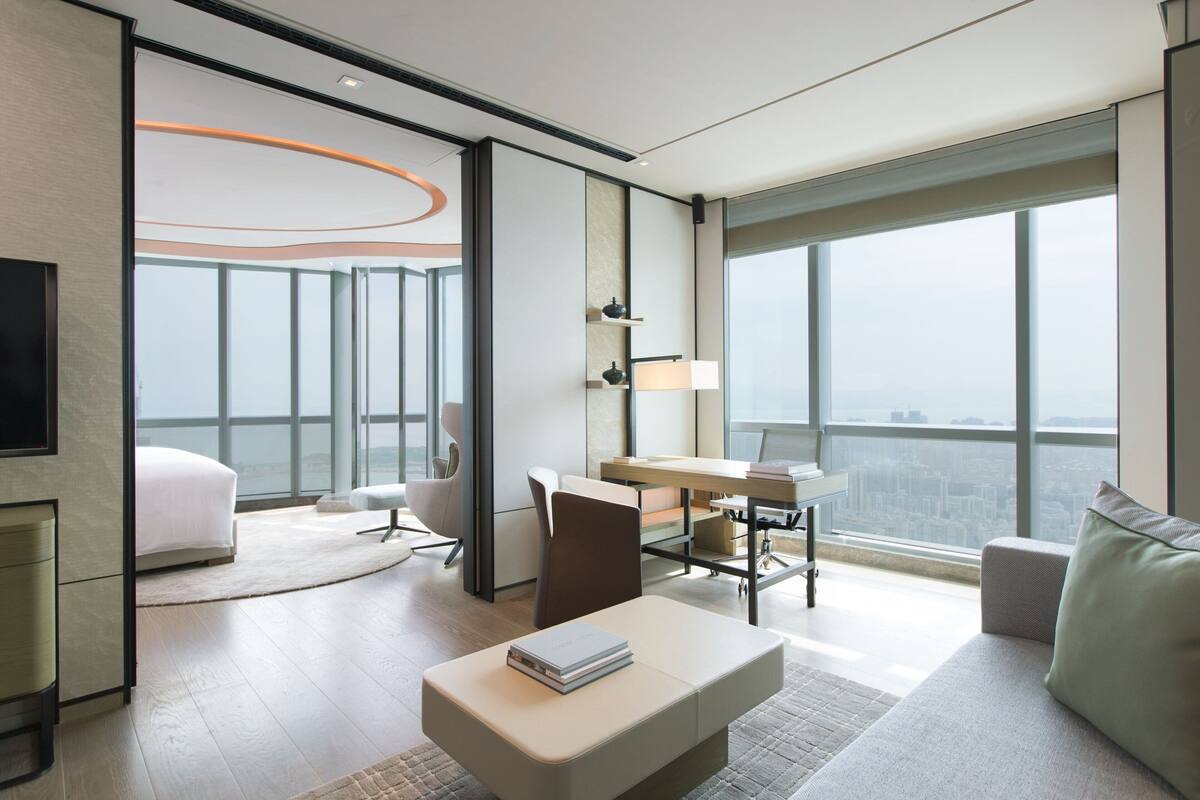 A modern hotel room with a large window, a desk, a chair, a sofa, and a flat-screen TV.