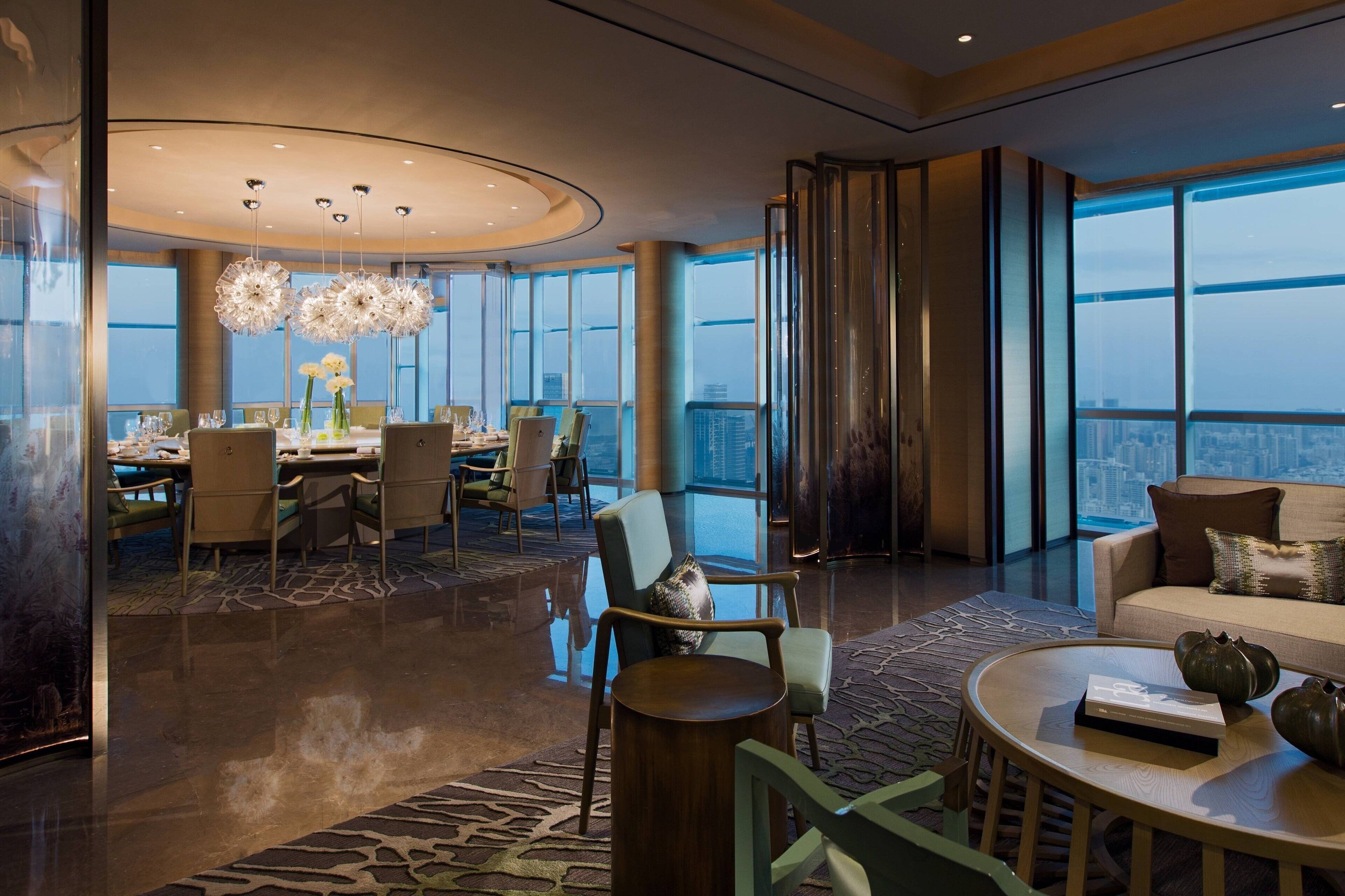 A modern hotel lobby with a high ceiling, large windows, and a dining area with round tables and chairs.