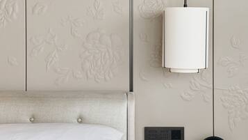 Een net opgemaakt bed met witte lakens, een houten nachtkastje met digitale klok en telefoon, en een muur met bloemversieringen.