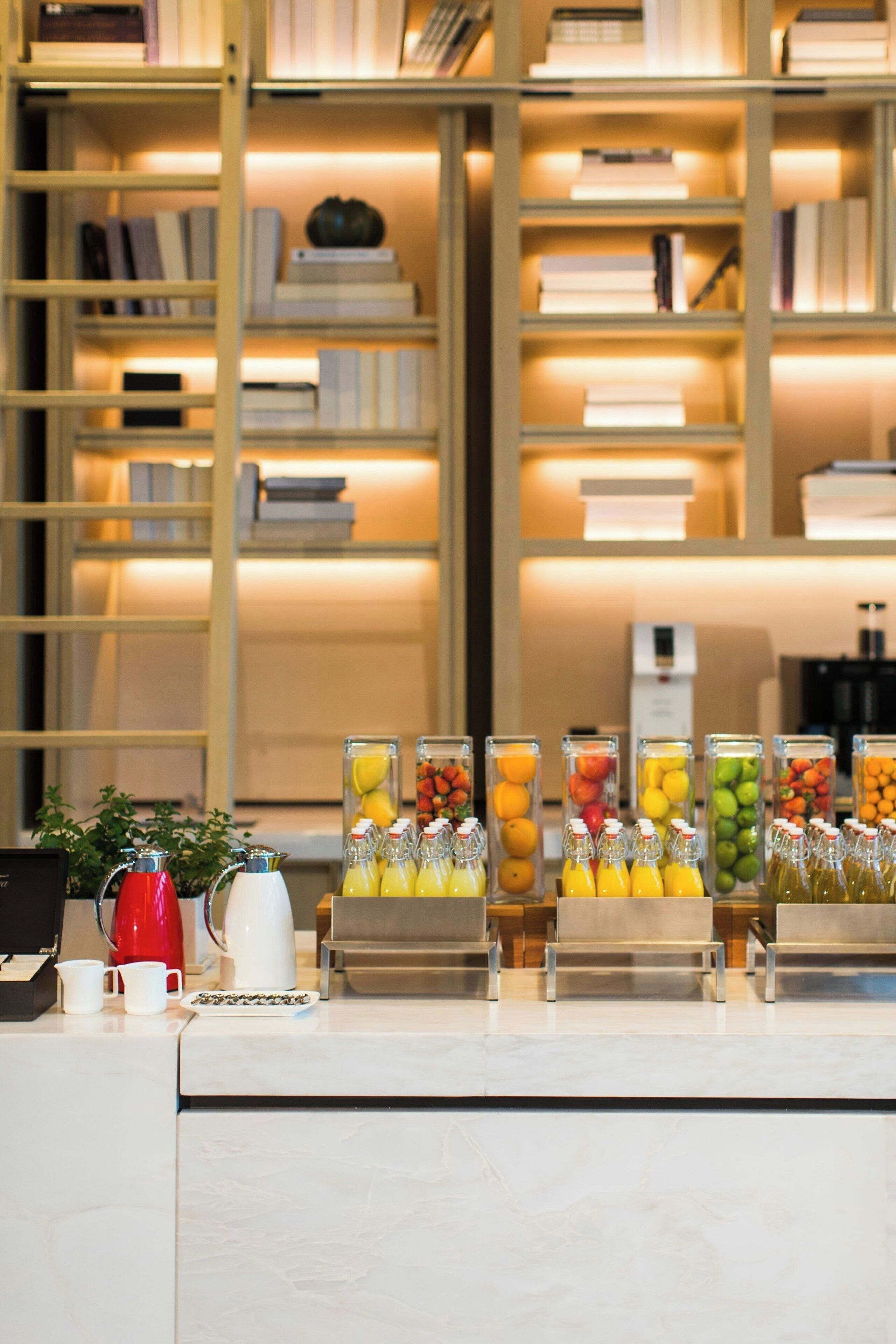 Zona de recepción con un mostrador que exhibe diversas bebidas en recipientes de vidrio, una cafetera y una pequeña planta. Detrás del mostrador, hay estanterías con libros y un electrodoméstico blanco.