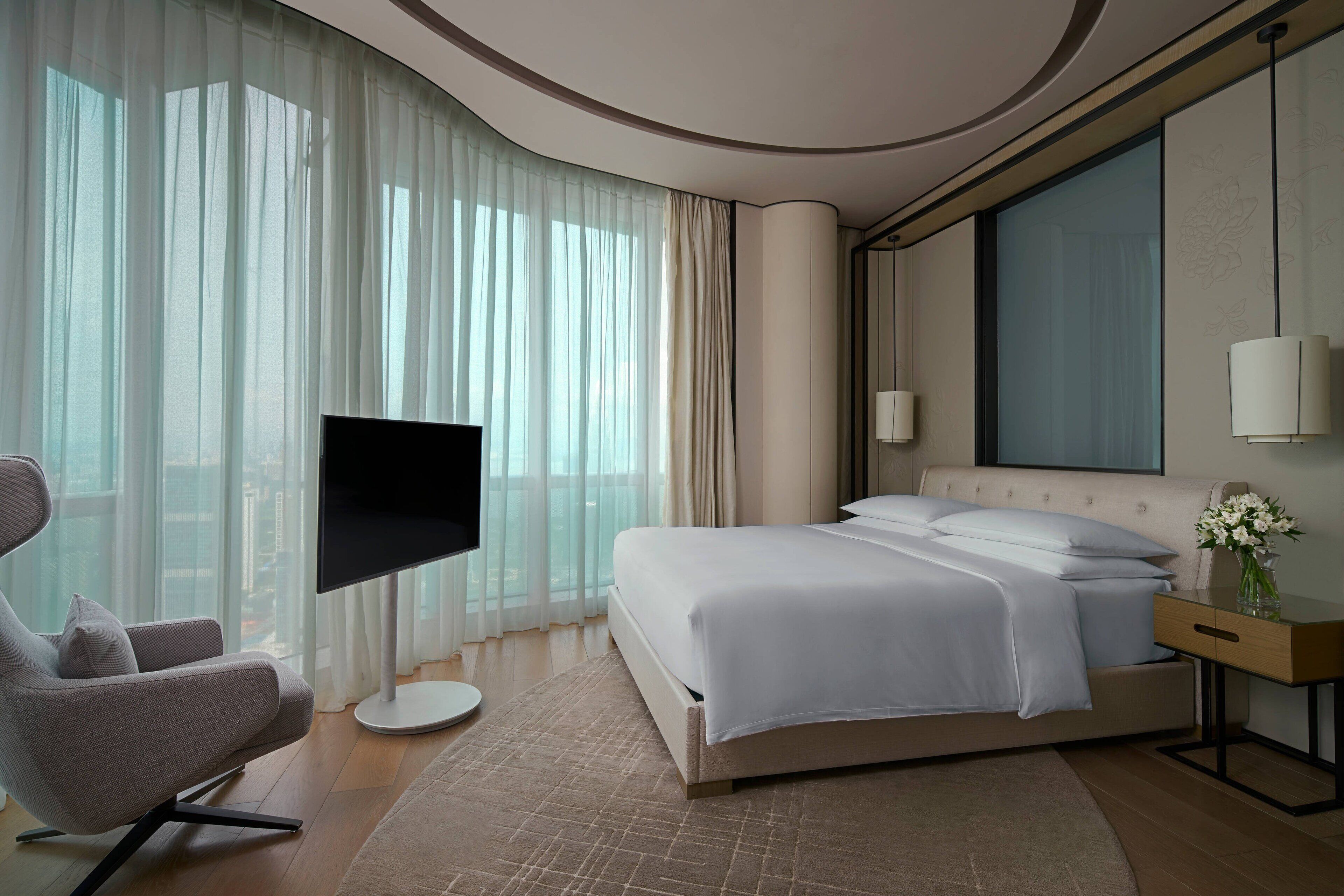 Una habitación de hotel moderna con una cama grande, un televisor de pantalla plana, un sillón cómodo y un ventanal con cortinas traslúcidas.