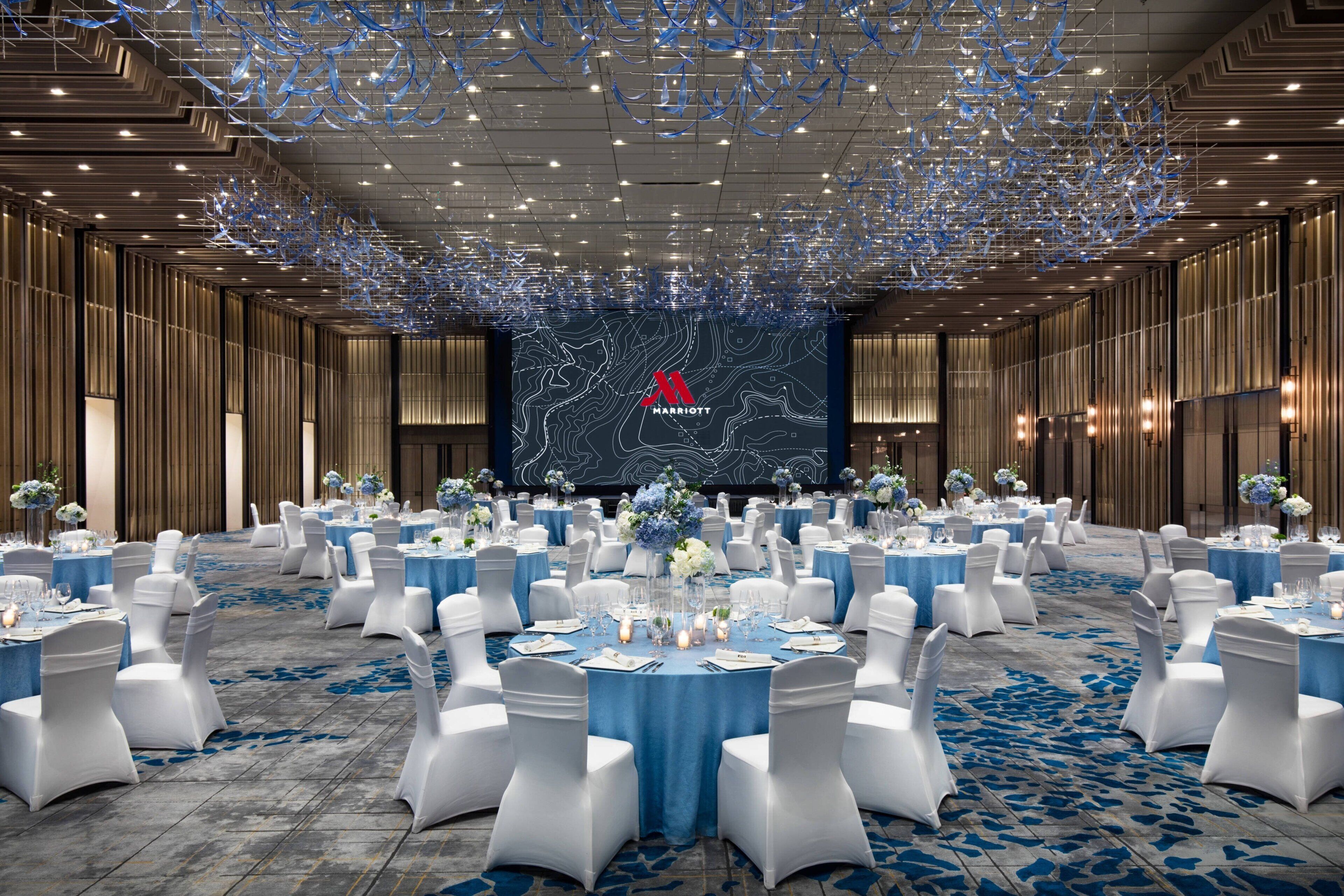 A large, well-lit banquet hall with round tables set for events, featuring a prominent Marriott logo on the back wall.