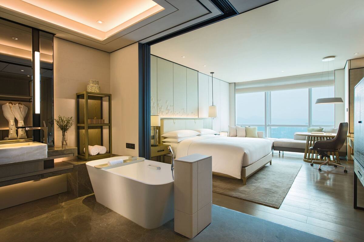 A modern hotel room with a large bed, a sitting area, a desk, a bathtub, and a view of the city.