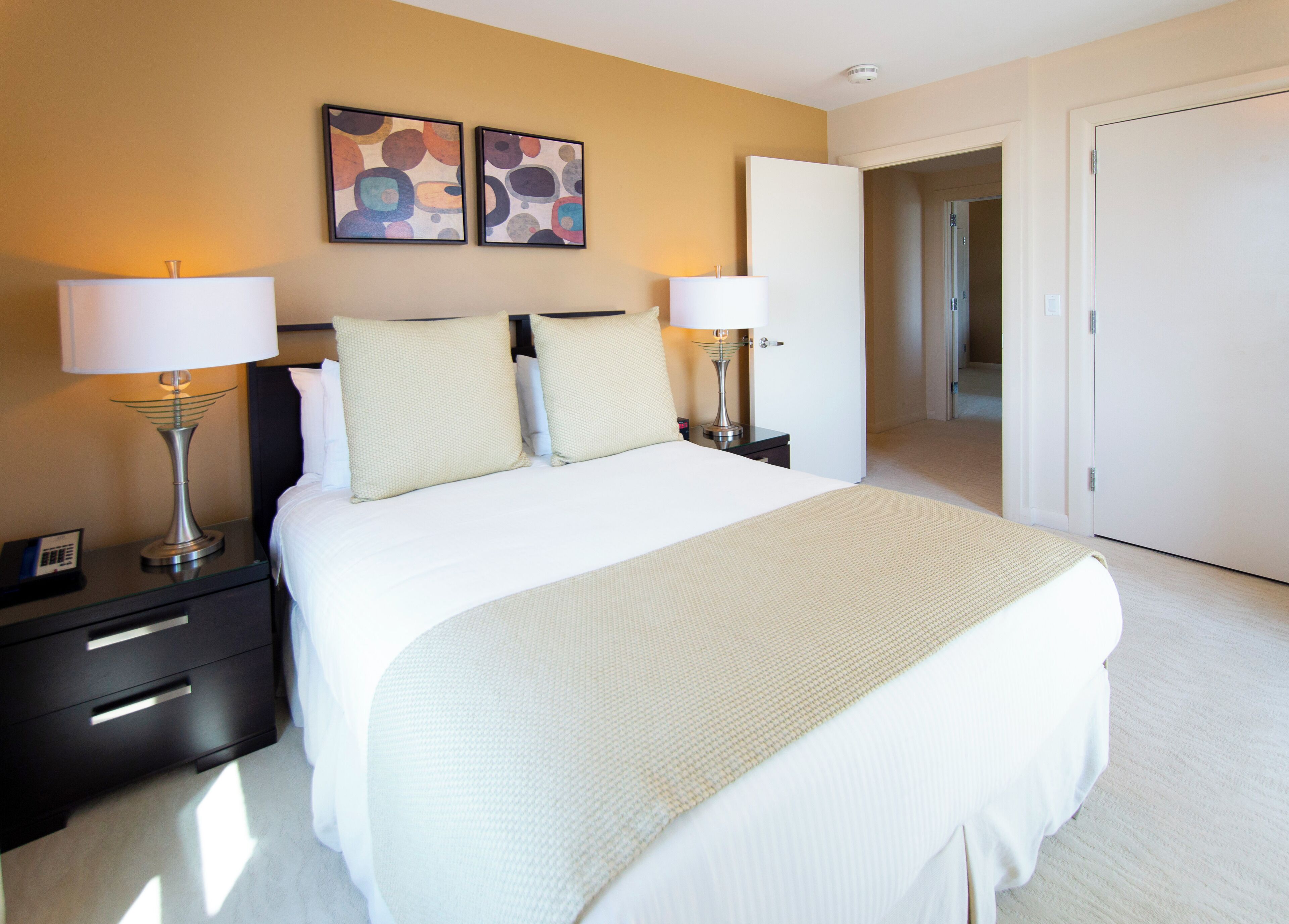 Une chambre d’hôtel avec un grand lit, deux tables de chevet avec des lampes, une table de nuit, une porte et des œuvres d’art abstraites accrochées au mur.
