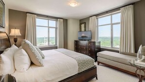 A bedroom with a large bed, a TV on a dresser, and a view of the city through large windows. - Broadway Plaza, Trademark Collection by Wyndham (Rochester)