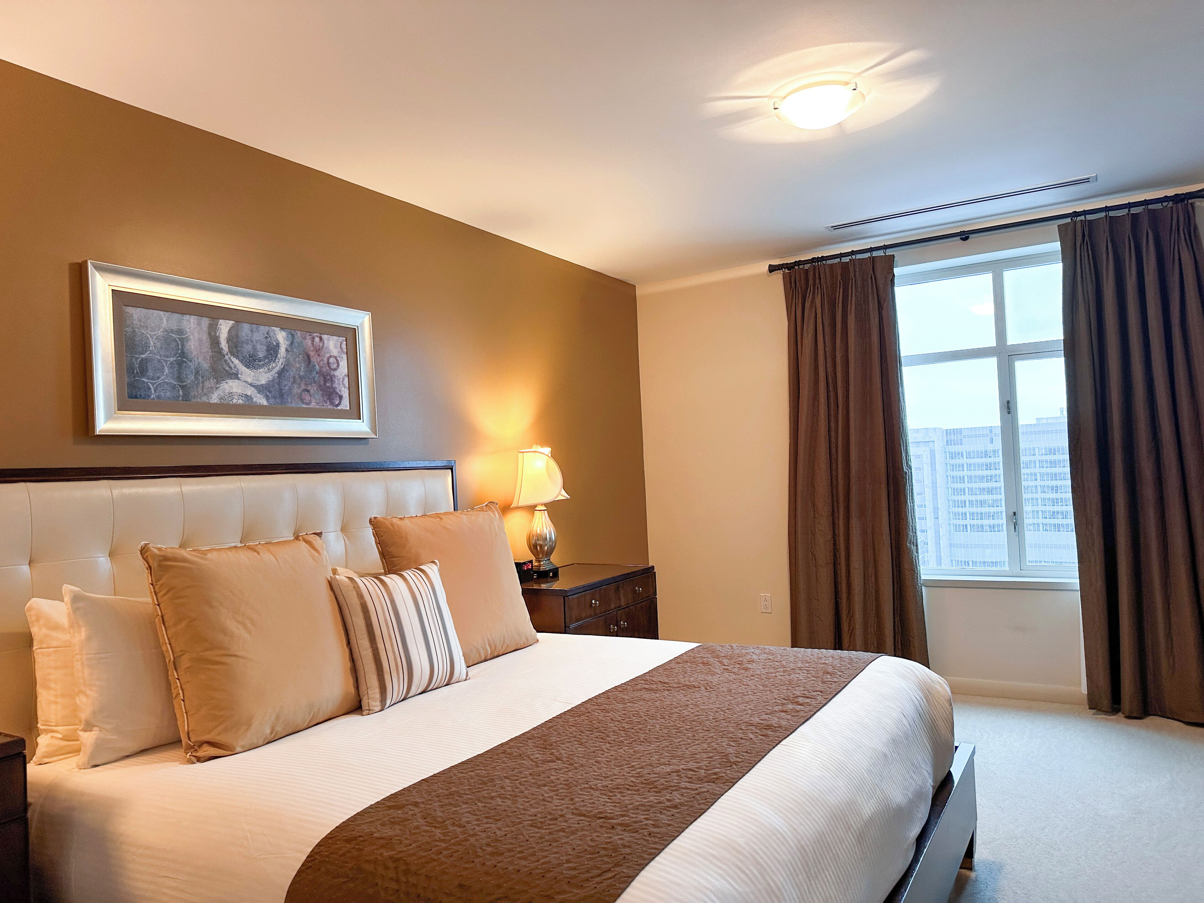 Ein Hotelzimmer mit einem großen Bett, einem Nachttisch mit Lampe, einem gerahmten Bild an der Wand und einem Fenster mit Vorhängen.