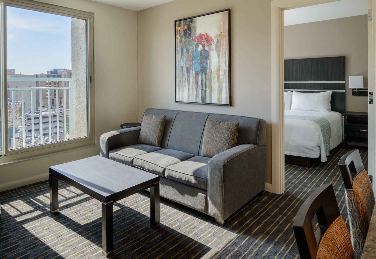 A hotel room with a gray sofa, a small table, a bed with white bedding, and a painting on the wall.