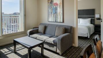 A hotel room with a gray sofa, a small table, a bed with white bedding, and a painting on the wall.