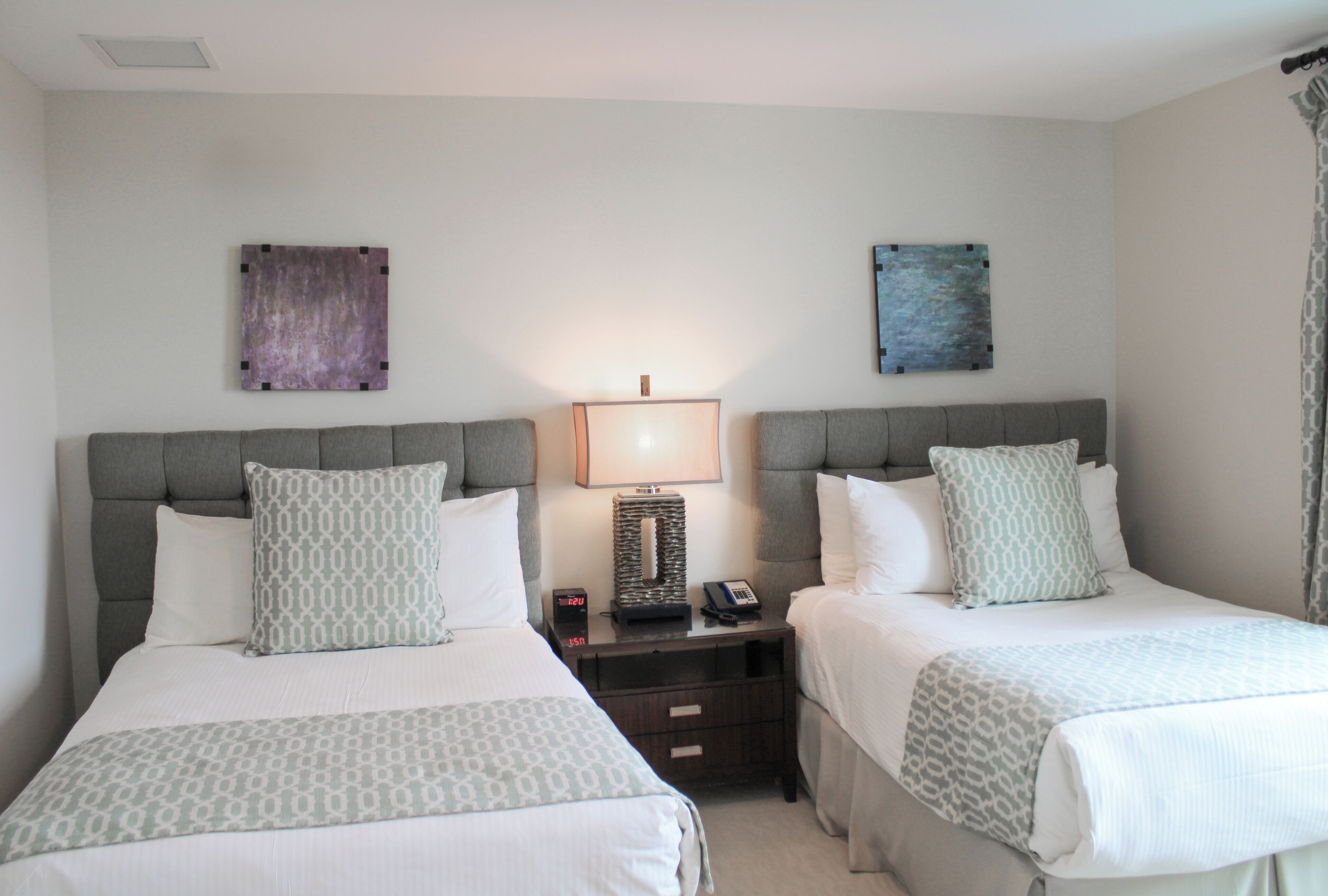 A hotel room with two beds, each with a grey headboard and white bedding. There is a bedside table with a lamp, a phone, and a small clock. The walls are plain with two pieces of abstract art.