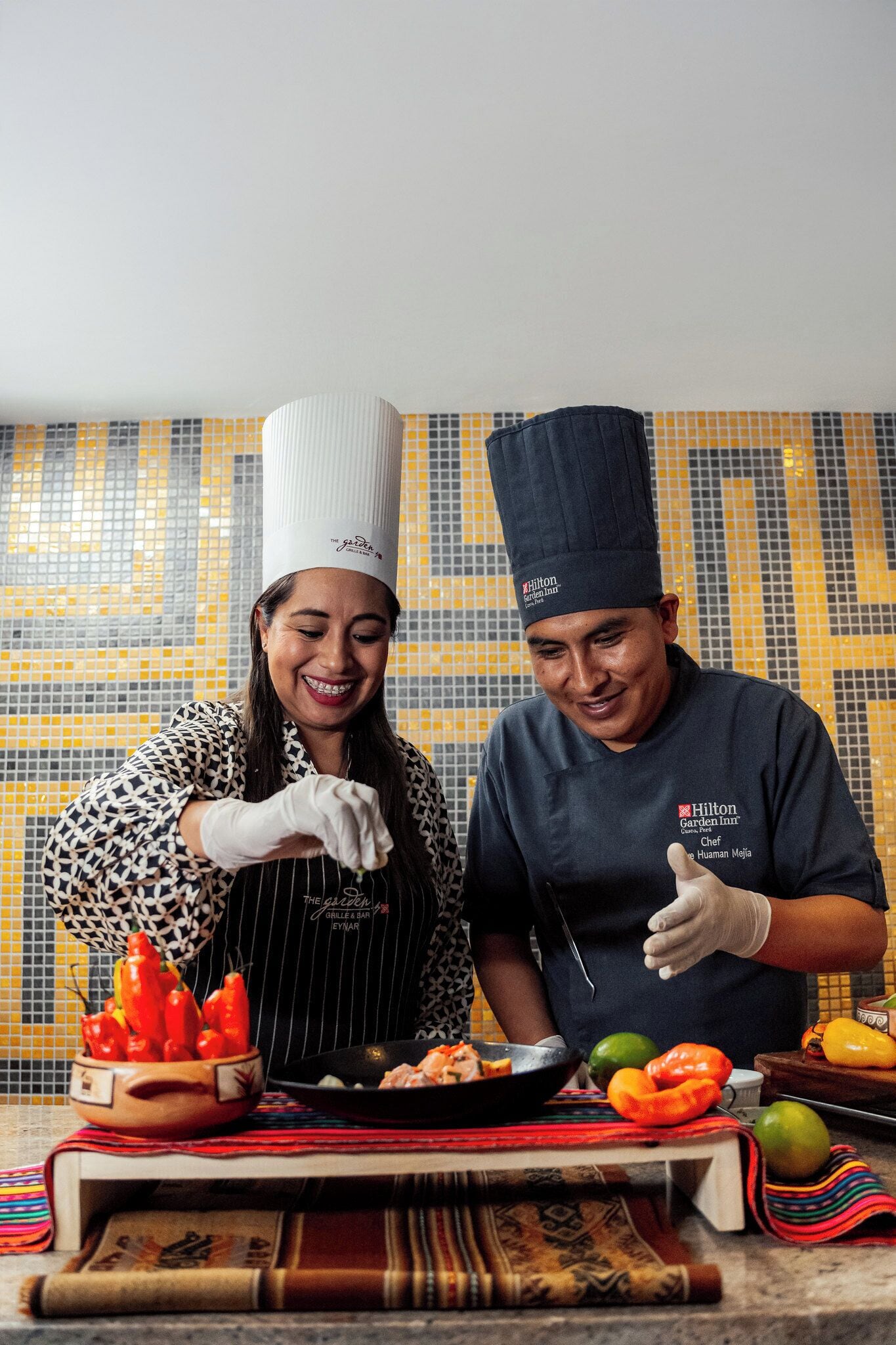 Dos cocineros en una cocina con un salpicadero de azulejos, un tazón de pimientos rojos y una sartén sobre la encimera.