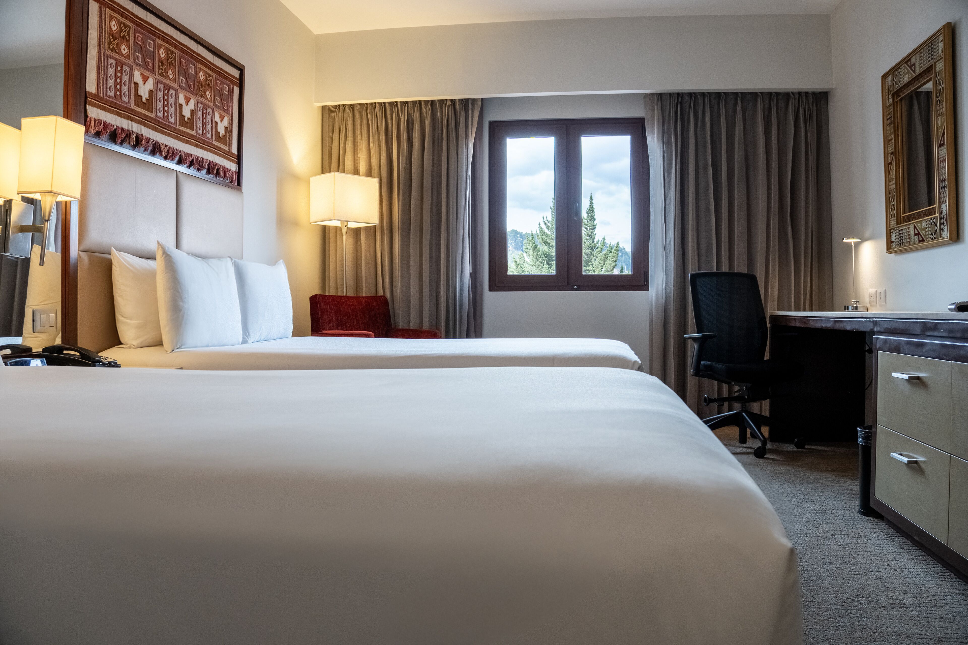 A hotel room with two beds, a desk, a chair, a lamp, and a window with curtains.