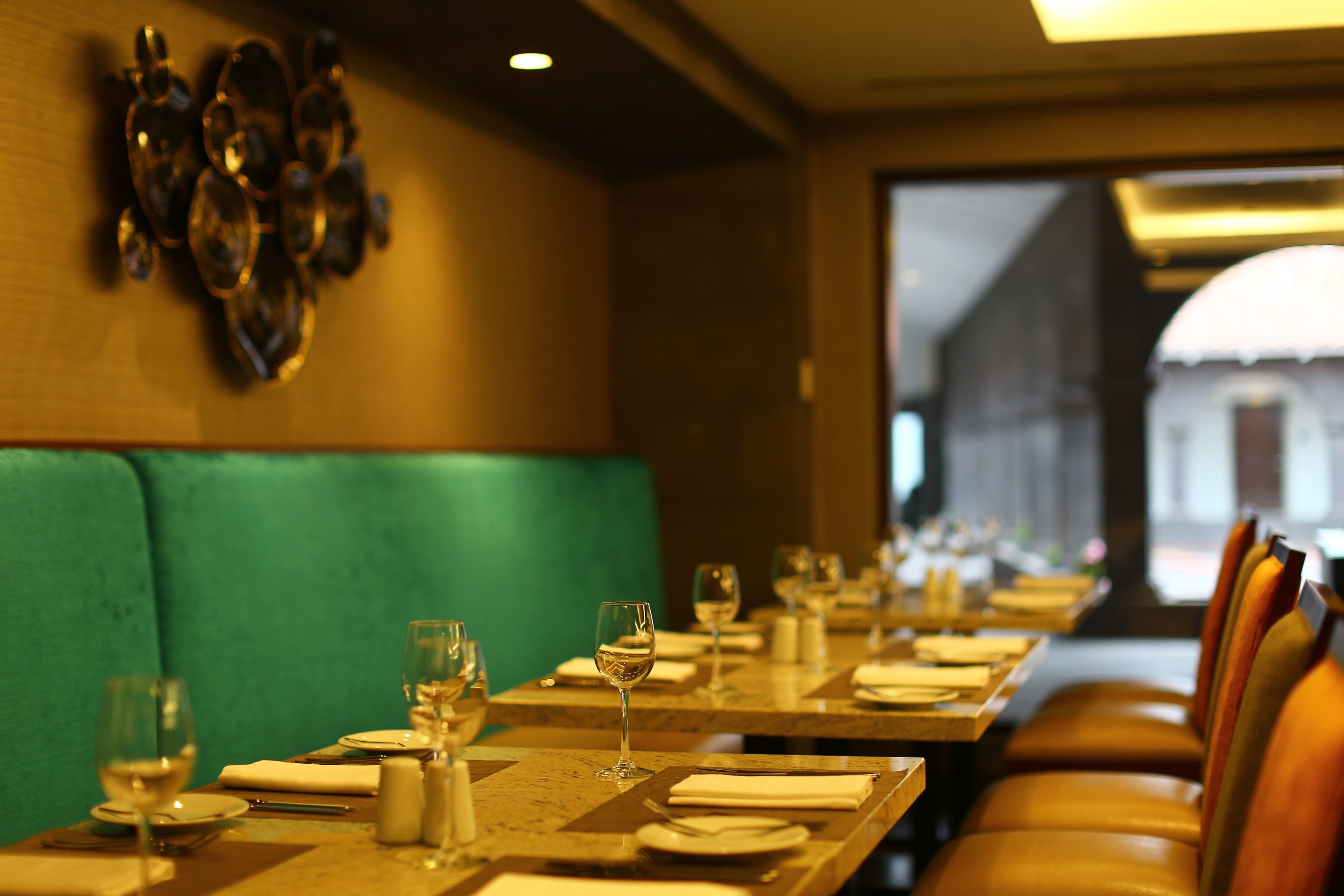 A restaurant with a long table set for dining, a green cushioned bench, and a view of a building through the window.