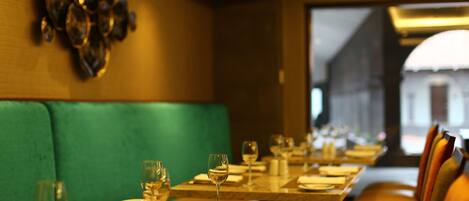 A restaurant with a long table set for dining, a green cushioned bench, and a view of a building through the window.